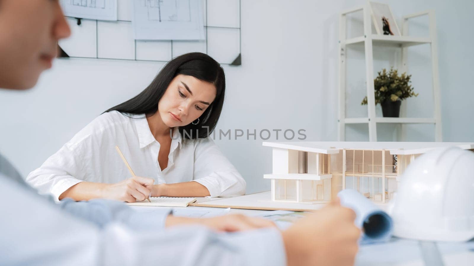 Professional architect engineer drawing blueprint and working together with young beautiful caucasian coworker on meeting table with safety helmet, house model, blueprint scatter around. Immaculate.