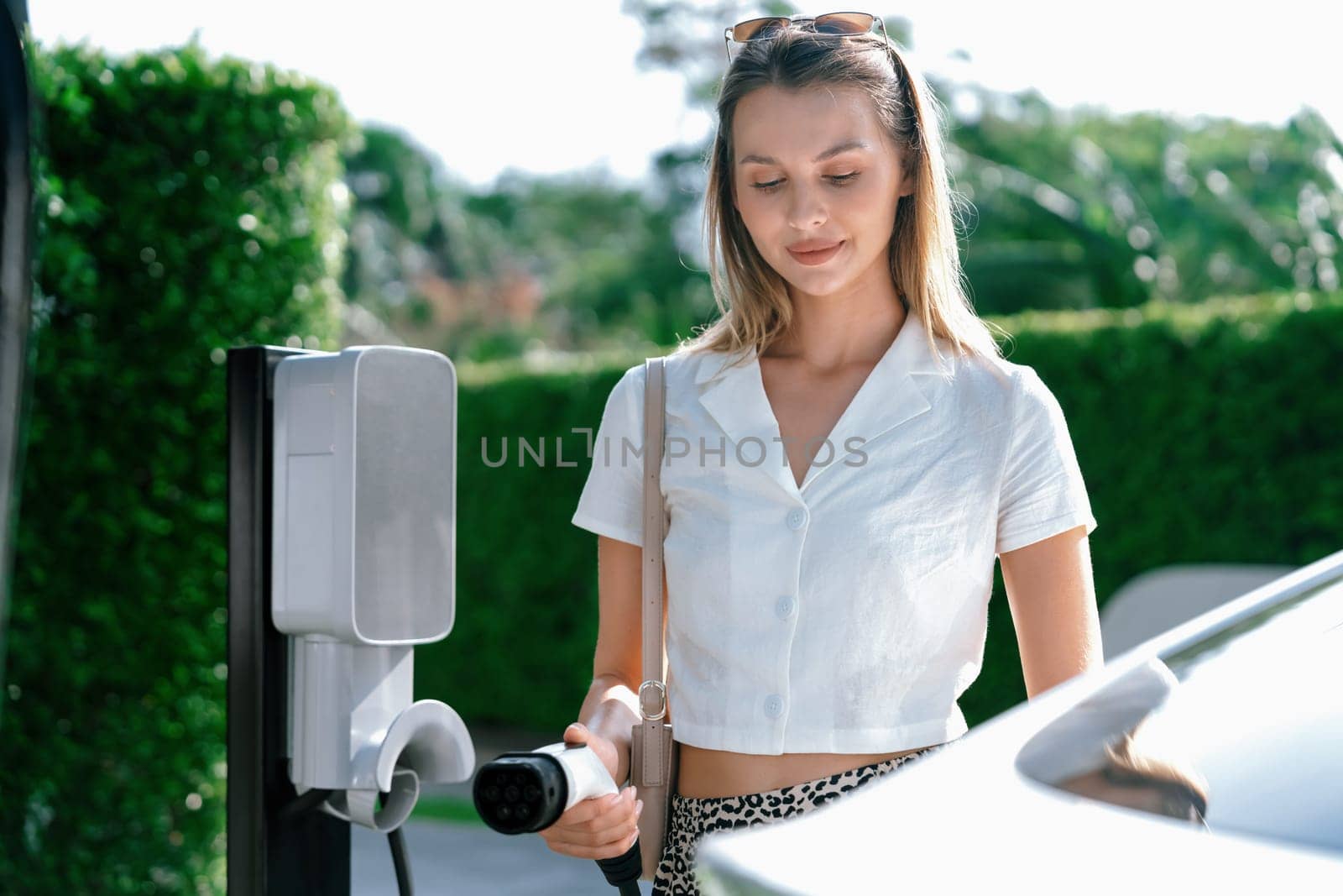 Young woman travel with EV electric car charging in green sustainable city outdoor garden in summer. Urban sustainability lifestyle by green clean rechargeable energy of electric BEV vehicle innards