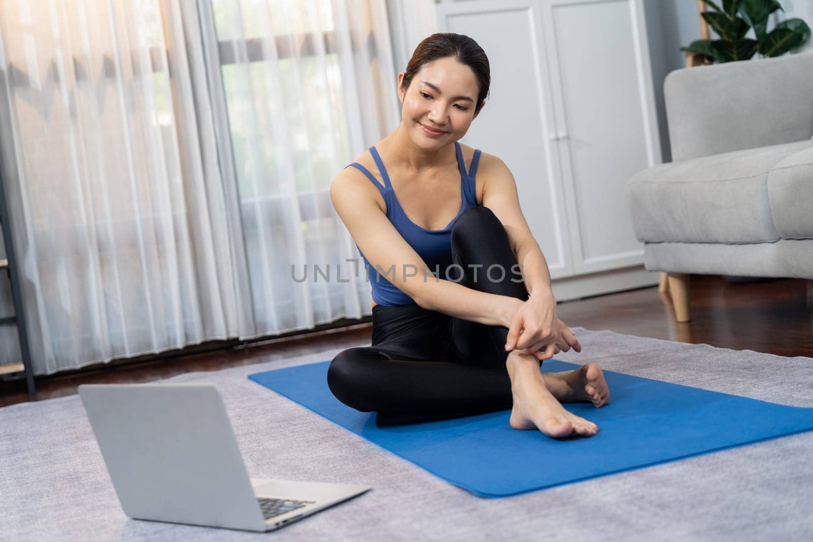 Asian woman in sportswear sitting on the house floor while watching online exercise training video on laptop. Attractive girl engage in her pursuit of healthy lifestyle. Vigorous