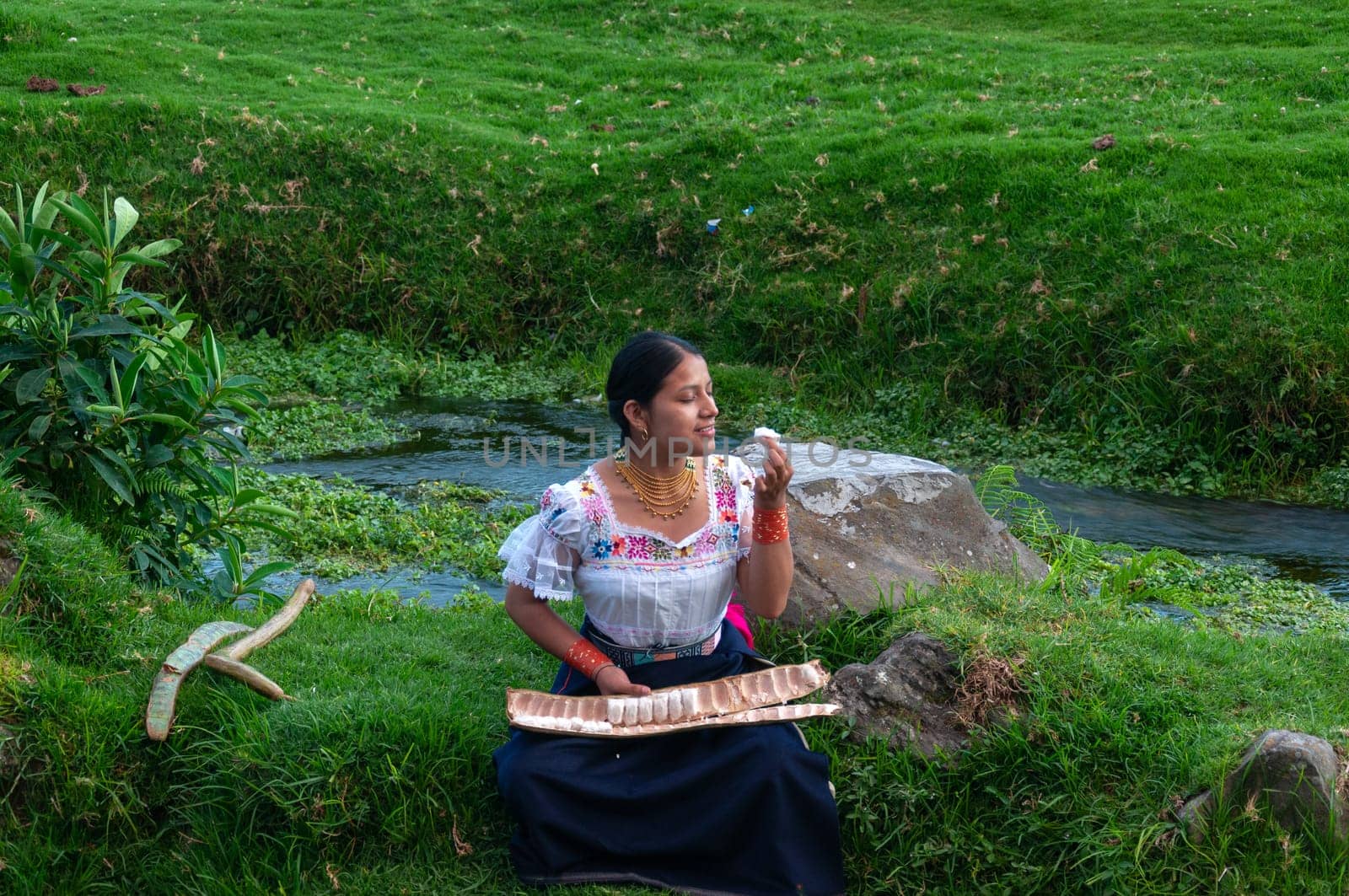 indigenous girl sitting next to a river tasting a large guaba, the girl wears a traditional dress from her culture. High quality photo