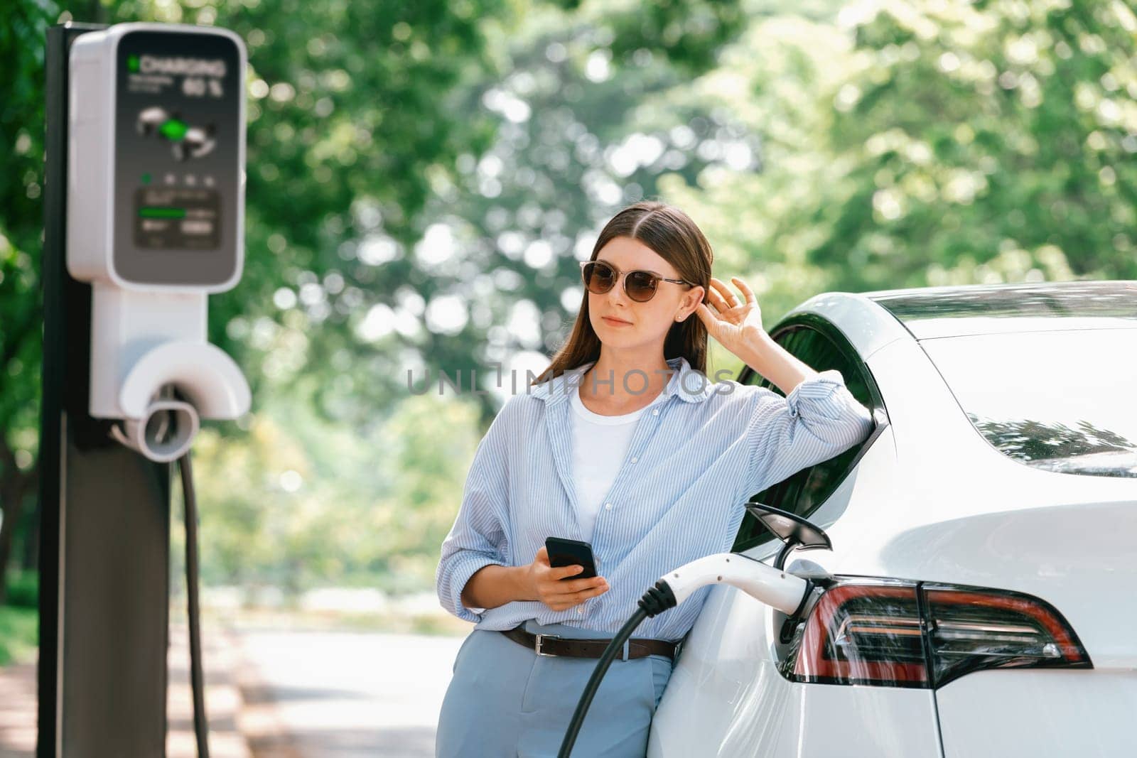 Young woman using smartphone online banking application to pay for electric car battery charging from EV charging station during vacation holiday road trip at national park or summer forest. Exalt