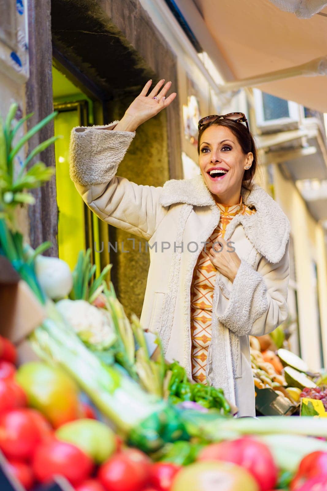 Cheerful woman waving hand while standing in fruits shop by javiindy