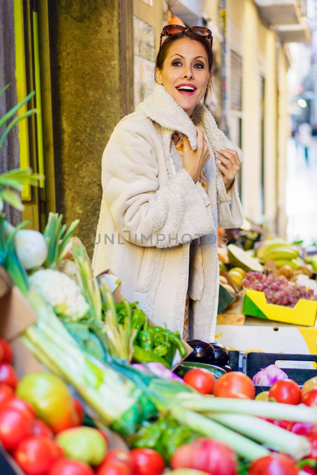 Side view of young woman in warm clothes, and eyeglasses on head looking at camera while standing in stall with assorted vegetables and fruits on sunny day