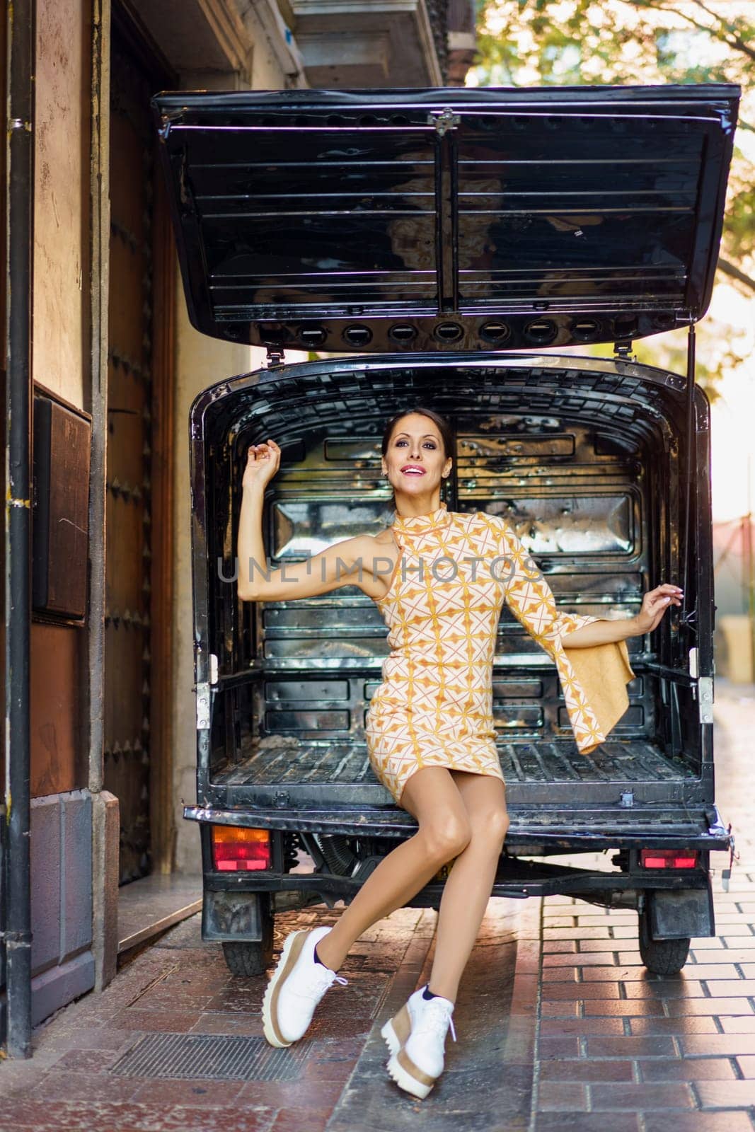 Full body of positive young female model, in stylish short gown and sneakers looking at camera and raising arms while sitting near opened door of caravan on street