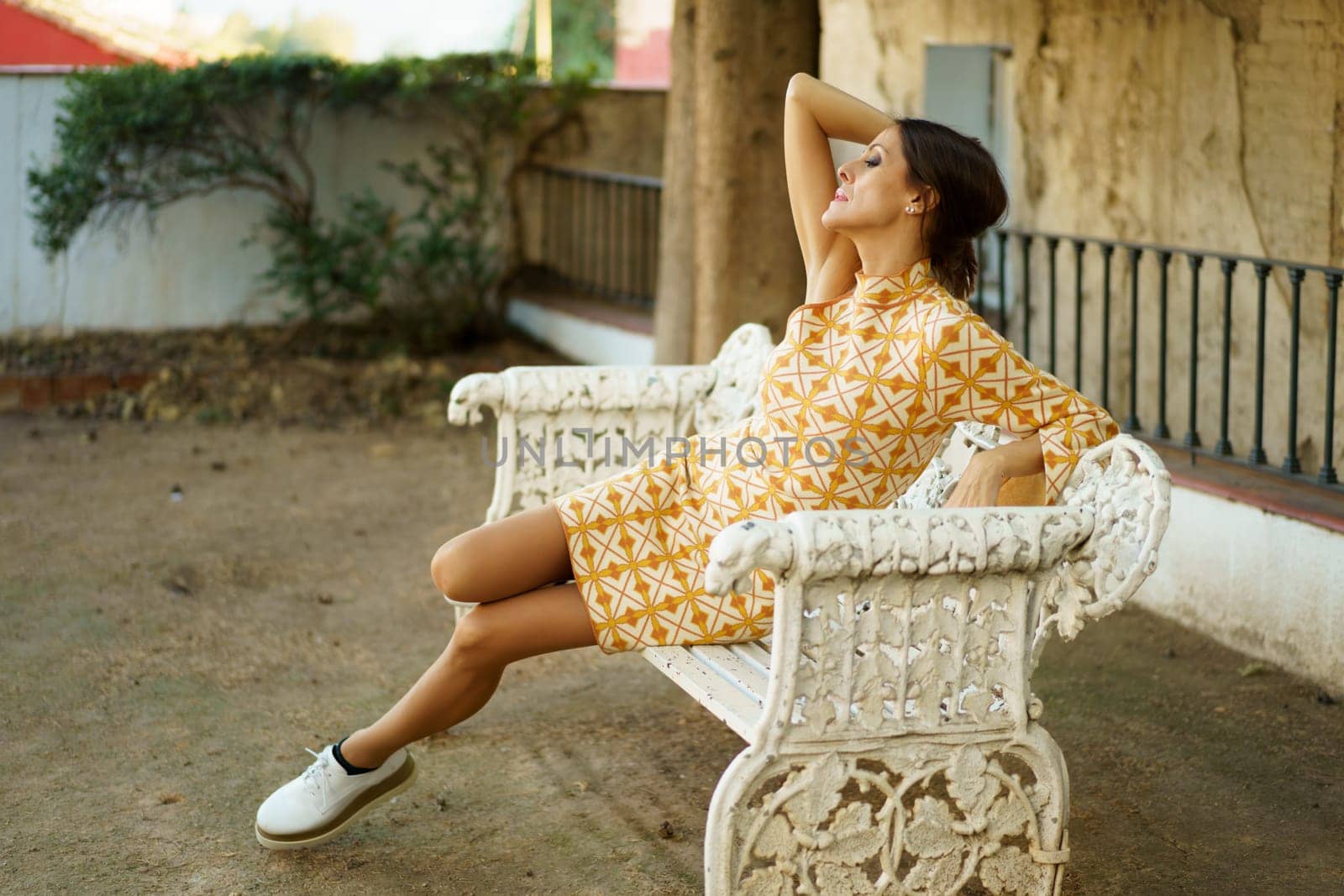 Full body side view of alluring young female model, in stylish dress and sneakers sitting with closed eyes and resting hand on backrest of bench by aged building