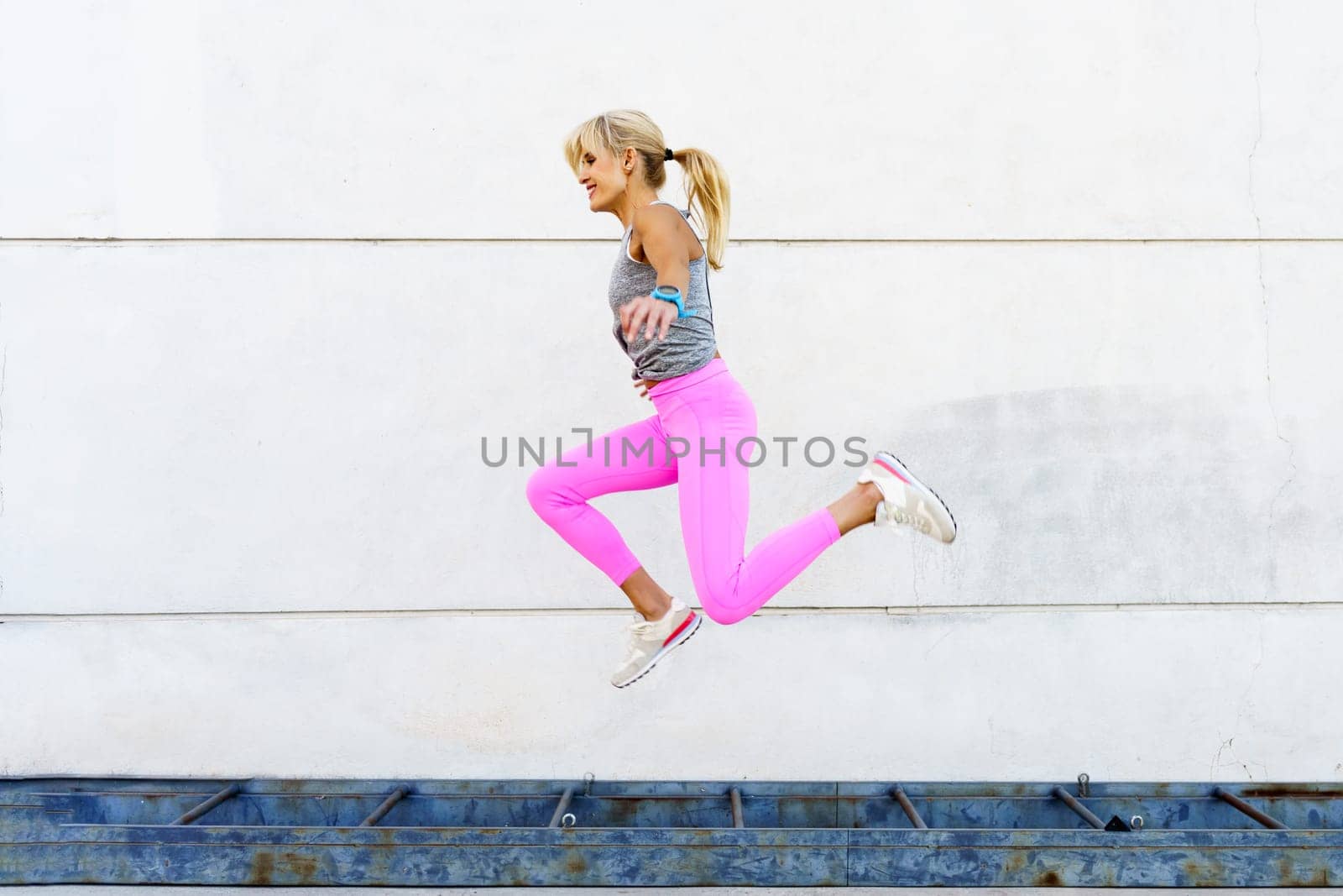 Side view of young female in activewear jumping above ground and smiling while training on street against white wall during fitness workout