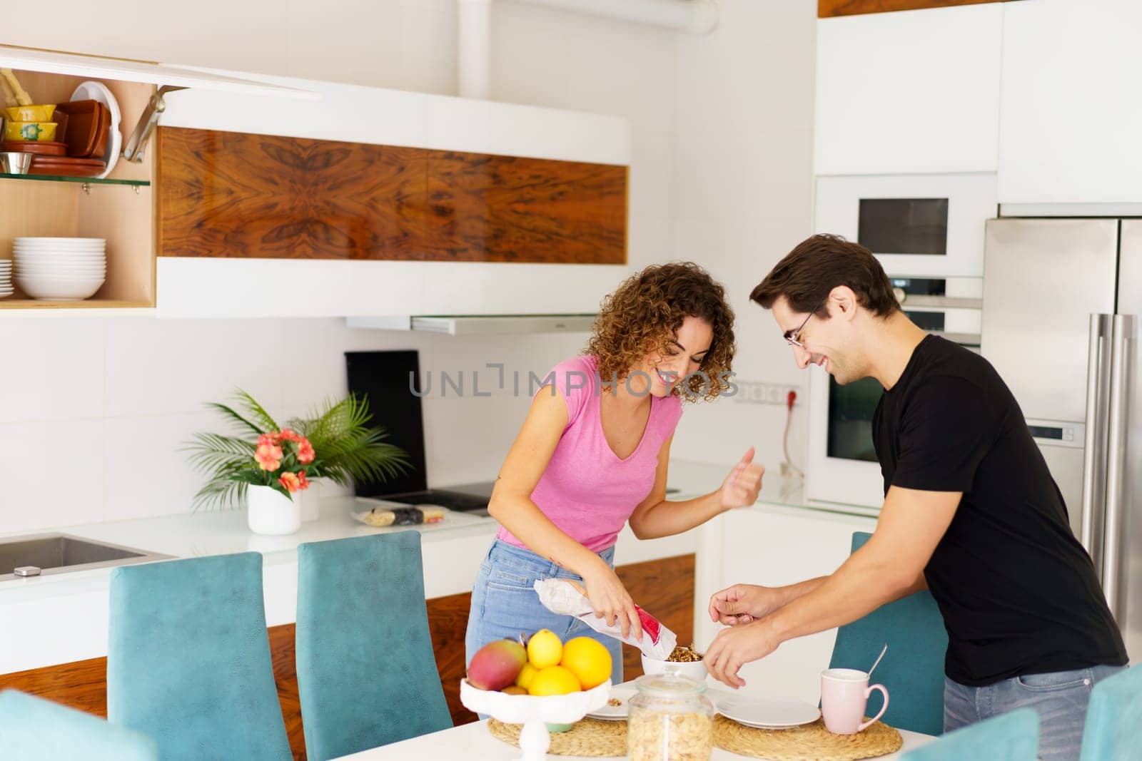 Side view of adult couple talking and looking down, while standing near dining table female pouring juice into glass and speaking with happy male in eyeglasses in kitchen in daylight