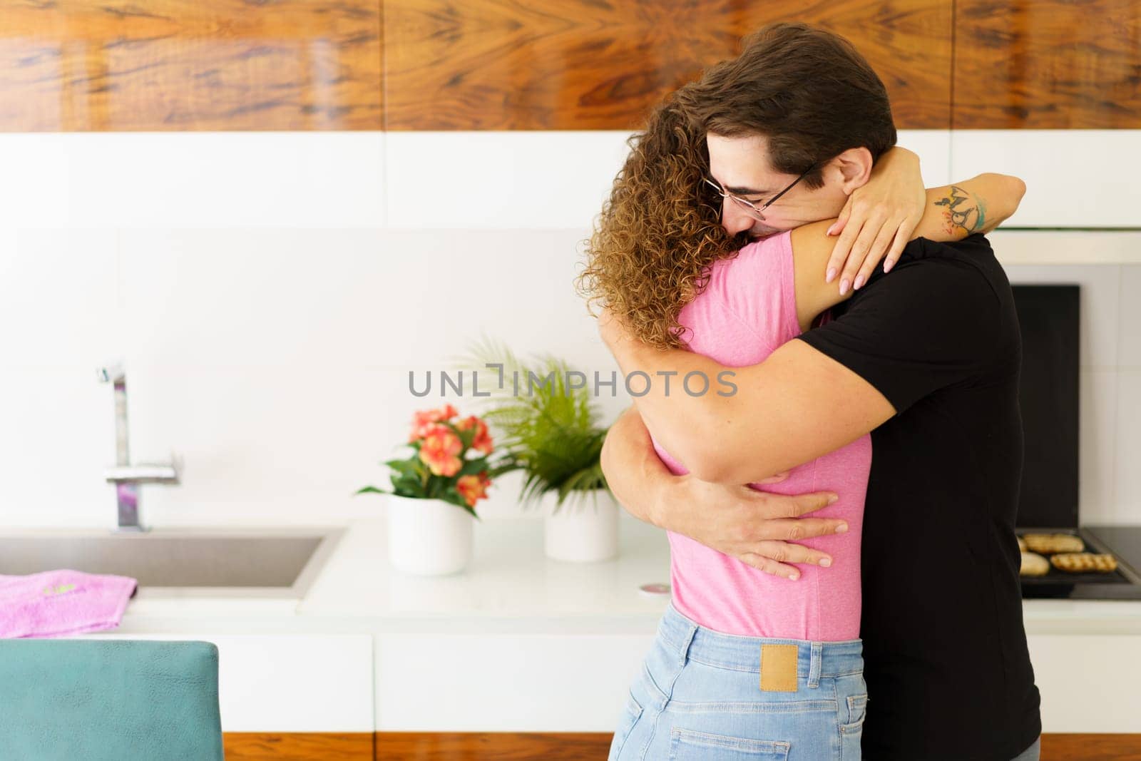 Side view of happy adult male in eyeglasses, looking down while standing in kitchen near cooking range with bread slices on grill and embracing faceless female with curly hair in daylight