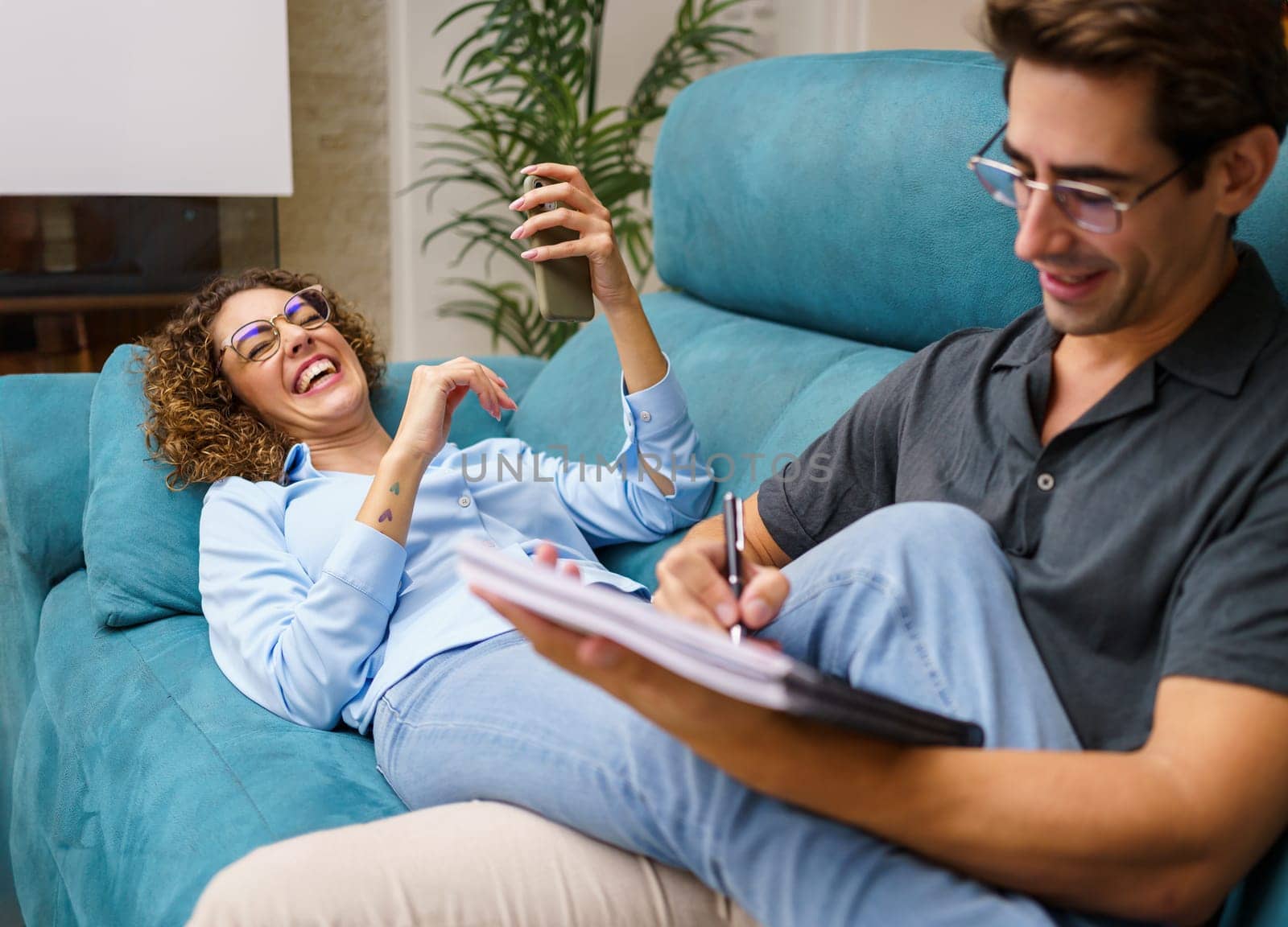 Happy young husband in casual clothes taking notes, while spending time with young curly hair wife in casual clothes laughing and lying with back on sofa in living room