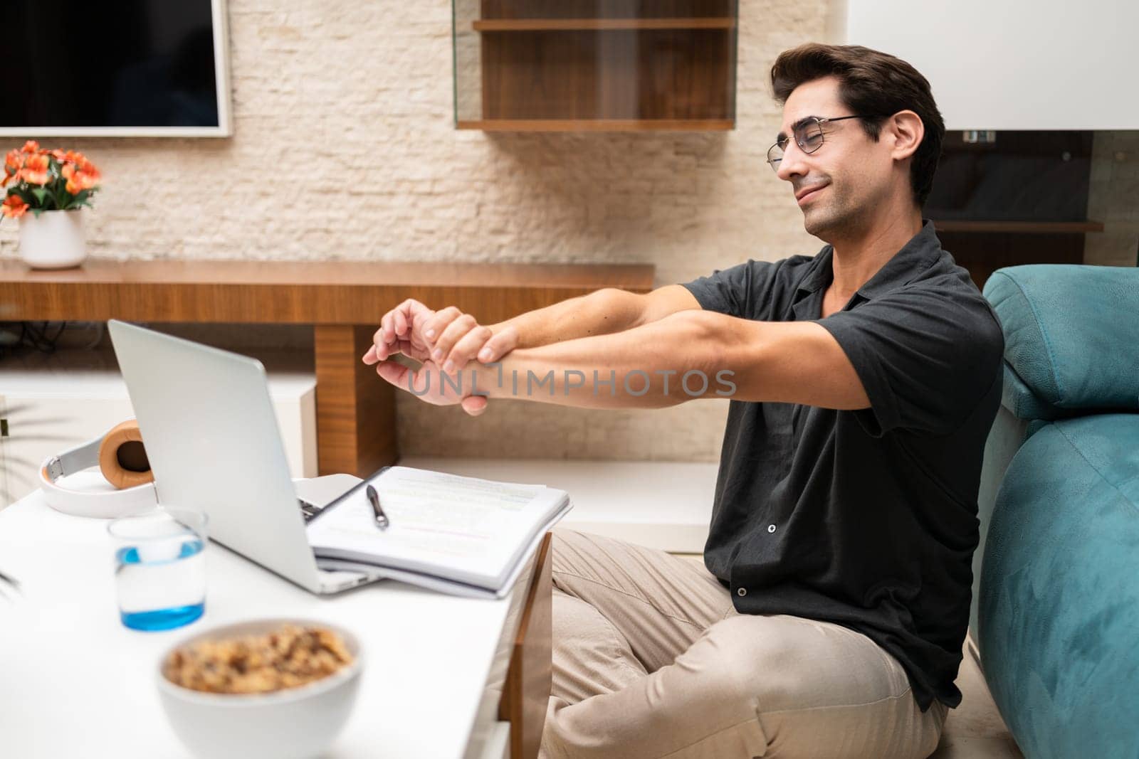 Side view of young male freelancer in casual clothes sitting near laptop and notebook while stretching arms at home
