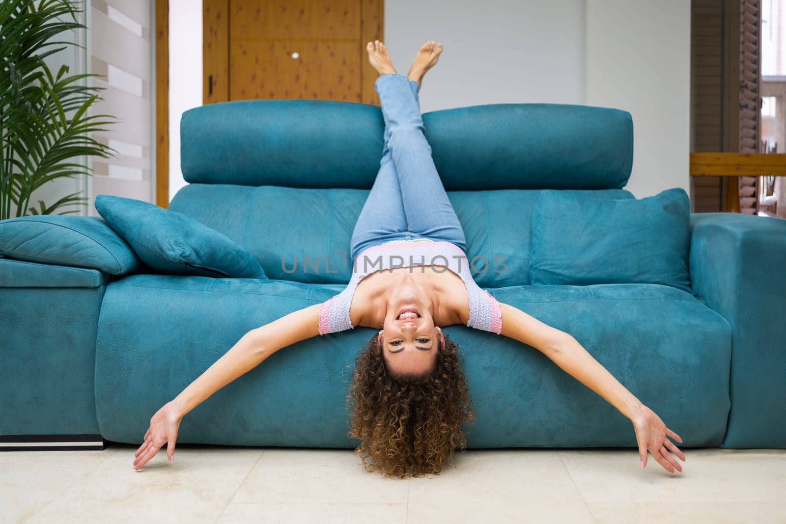 Smiling young barefoot female with curly hair lying upside down on couch and looking at camera while having fun at home
