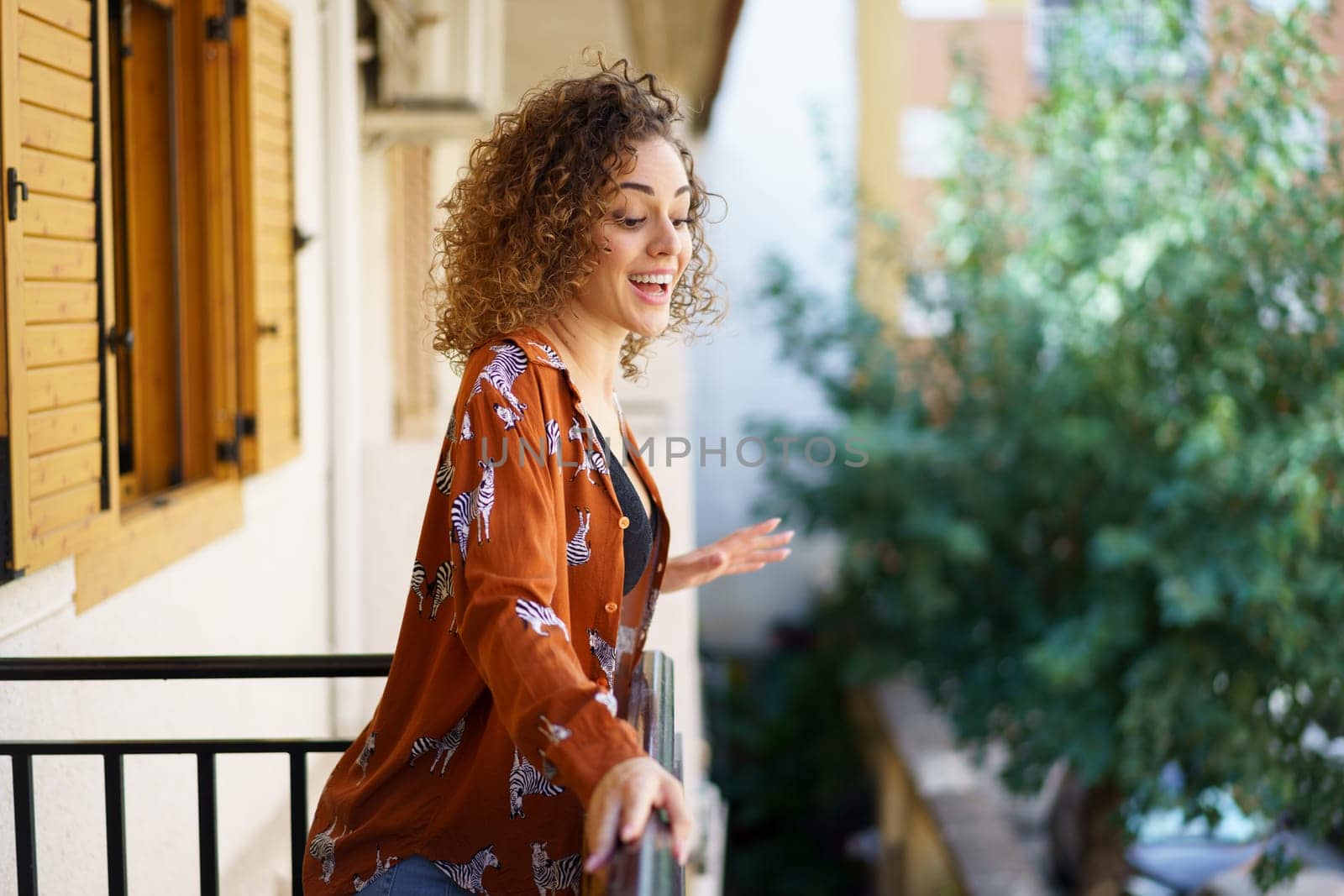 Joyful young brunette curly hair woman standing on balcony of modern building by javiindy