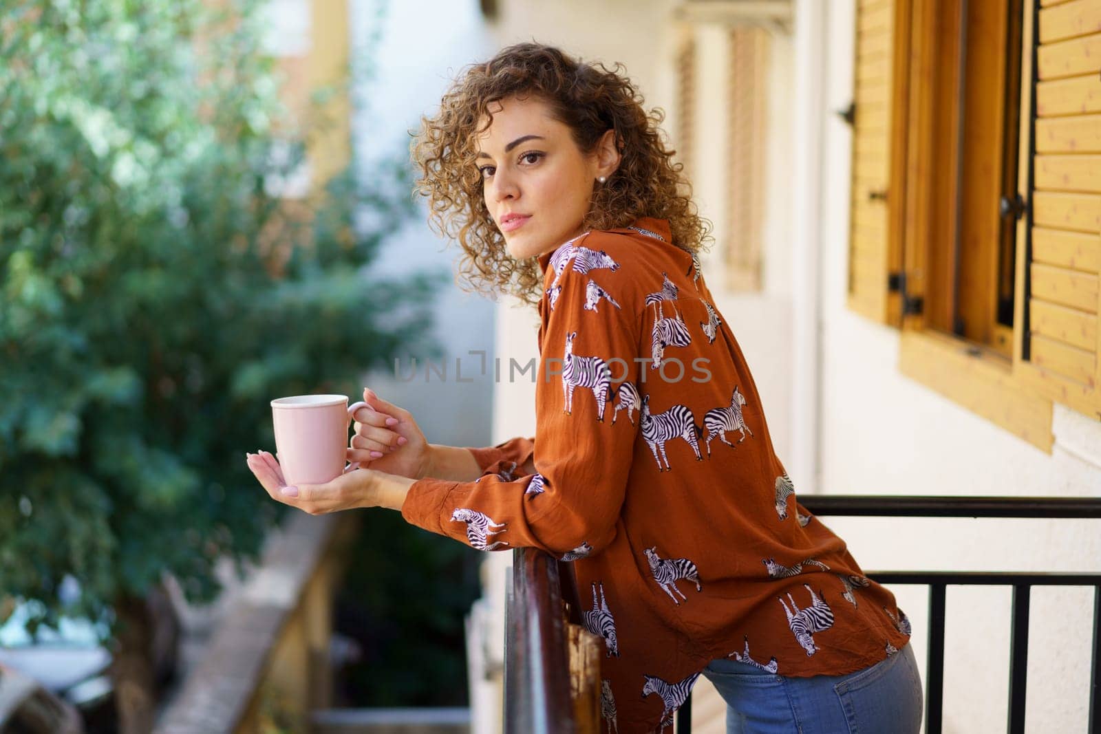 Side view of young female wearing casual clothes leaning on balcony railing while looking away and holding coffee cup against blurred background
