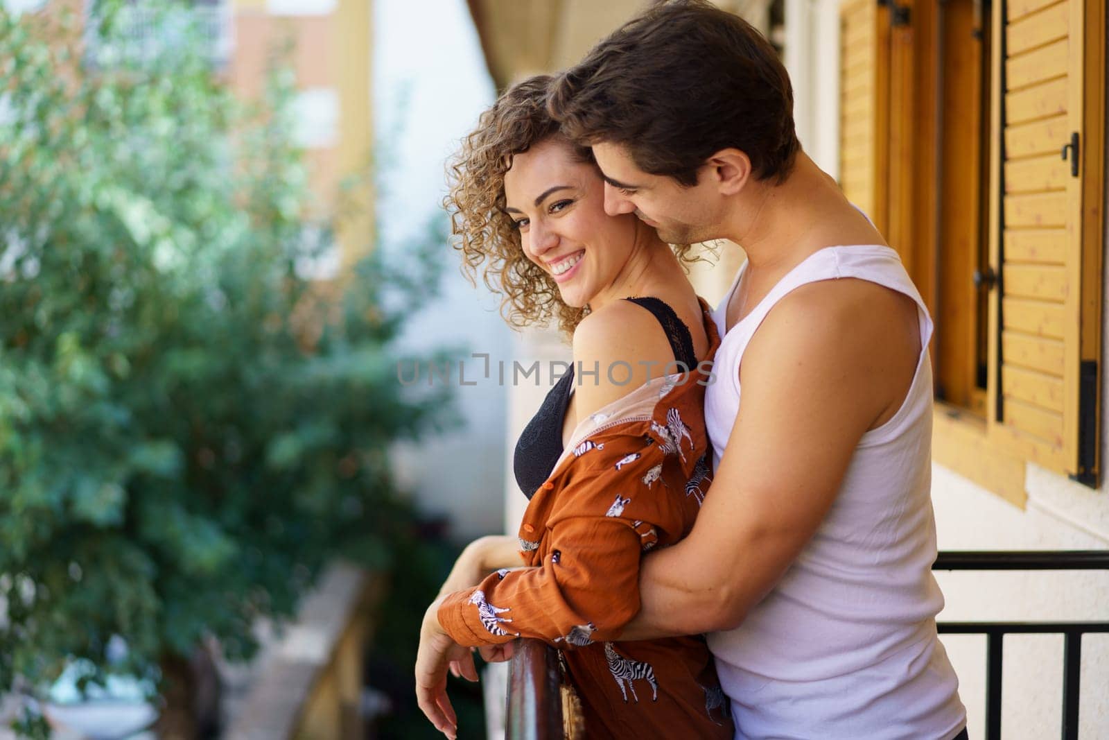 Side view of loving cheerful young couple in inner wears, embracing and hugging while standing by rail on balcony of apartment over blurred nature background on sunny day