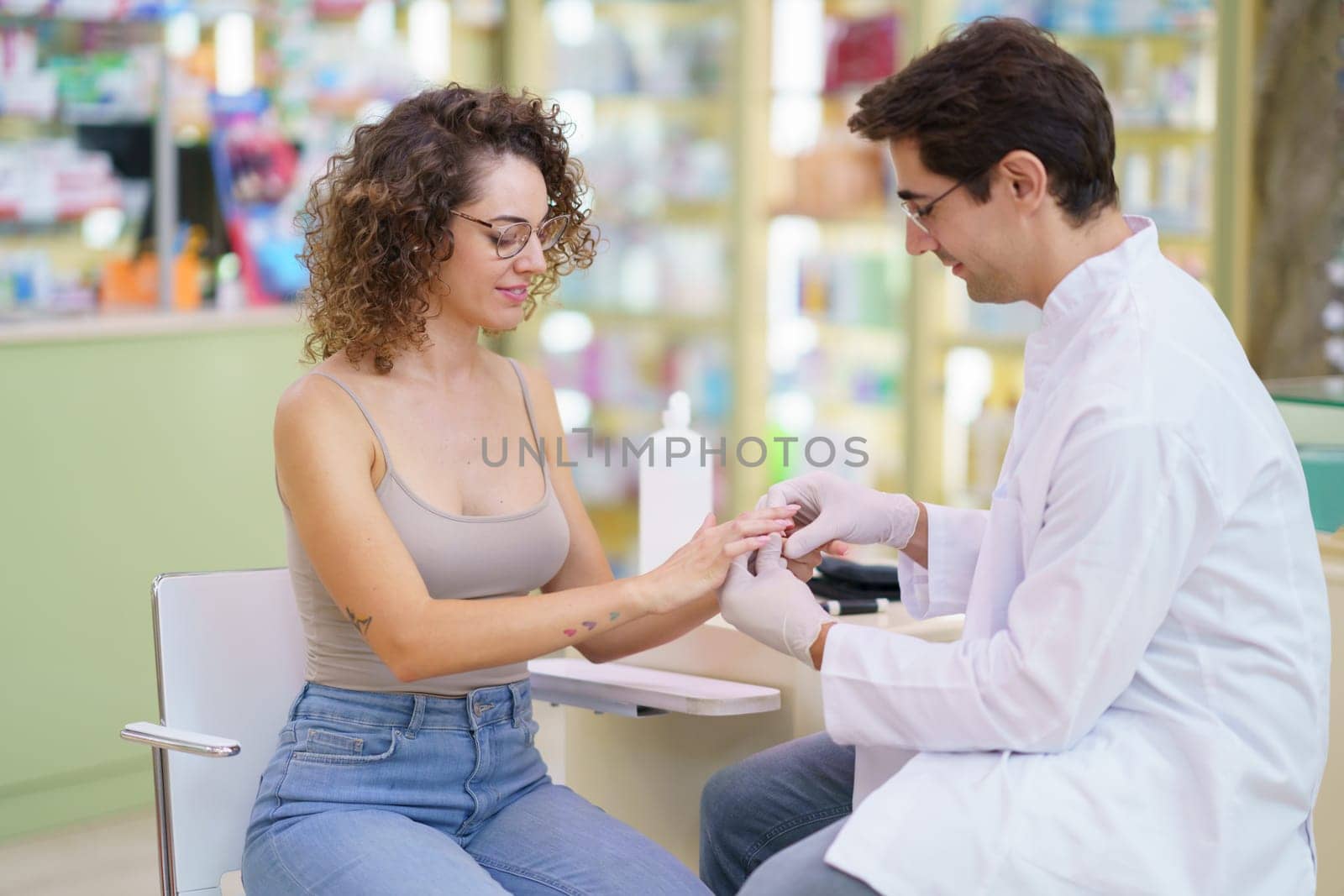 Male doctor in white uniform checking blood sugar level of female patient sitting in pharmacy