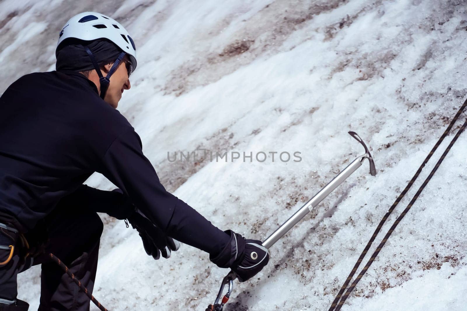 A young man traveler uses an ice ax while hiking in the mountains, a hiker with climbing equipment, in a helmet, crampons climbs to the top of a snow-capped mountain.