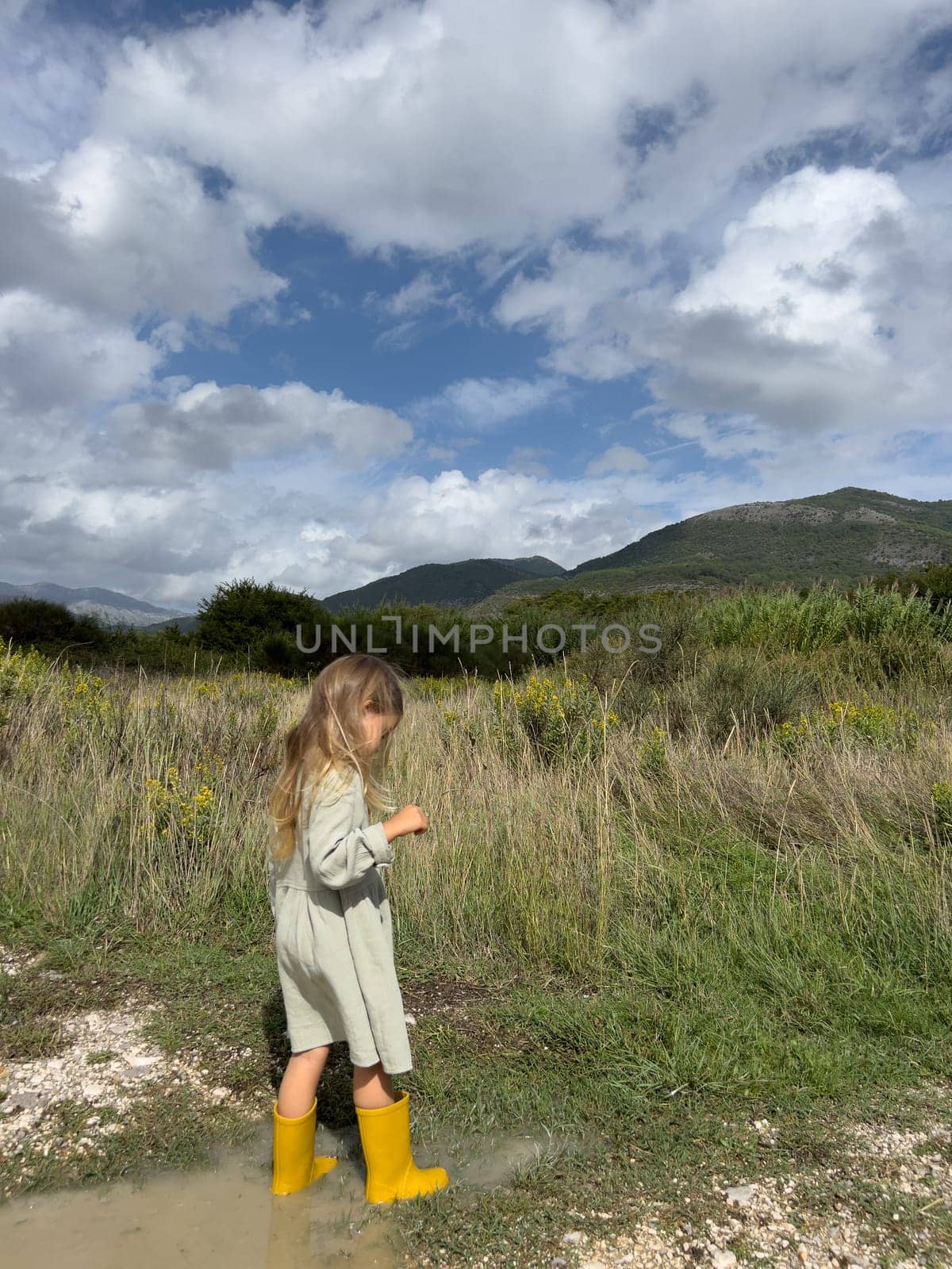 Little girl in rubber boots walks through puddles in a mountain valley by Nadtochiy