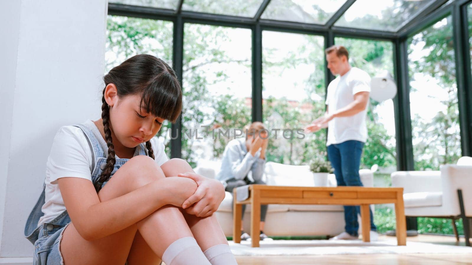 Stressed and unhappy young girl huddle in corner, cover her ears blocking sound of her parent arguing in background. Domestic violence at home and traumatic childhood develop to depression. Synchronos