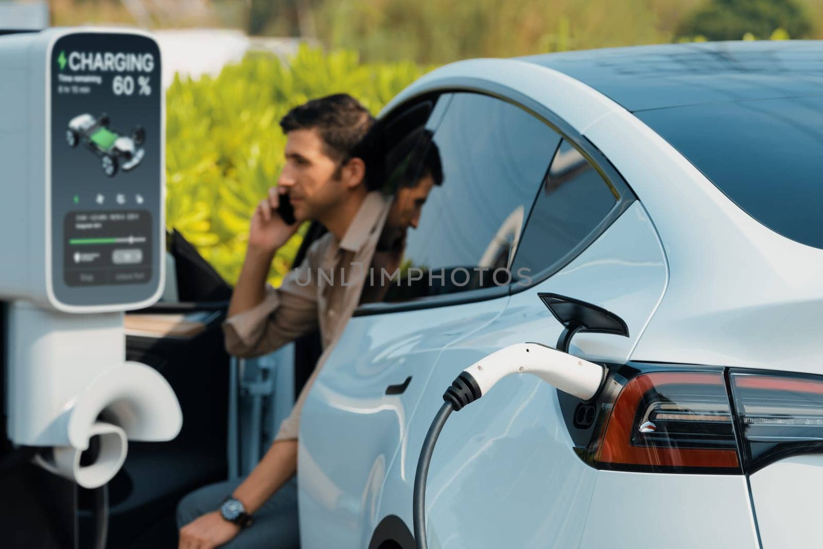 Young man recharge EV electric vehicle at green city commercial mall parking lot while talking on phone. Sustainable urban lifestyle for eco friendly EV car with battery charging station. Expedient