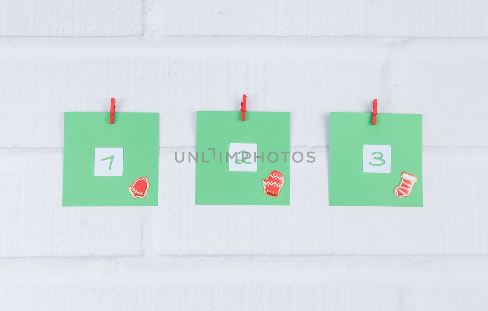 Three green sheets of paper with numbers 1,2,3 and Christmas decor are attached with red mini clothespins on a white brick wall, side view close-up. Concept diy advent calendar, creative and minimalism.