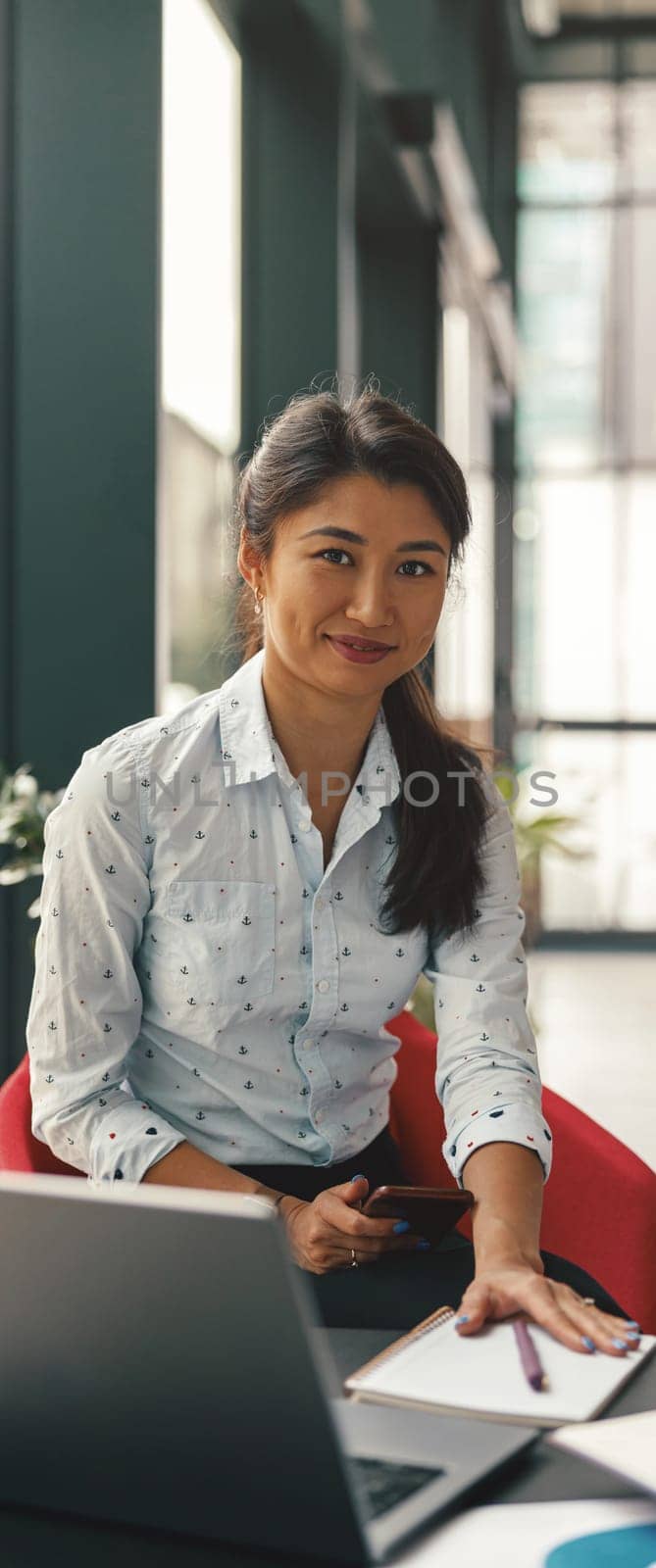 Smiling business woman holding phone and looking at camera while working on laptop in office