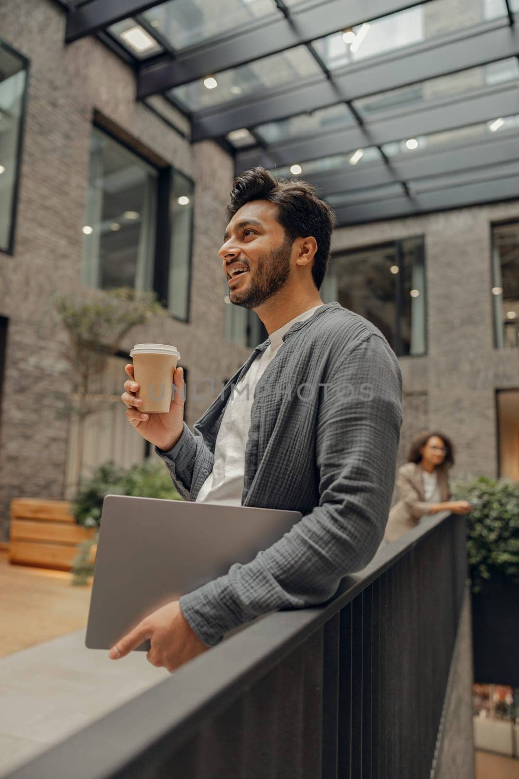 Handsome businessman with laptop is standing on office background and drinking coffee by Yaroslav_astakhov