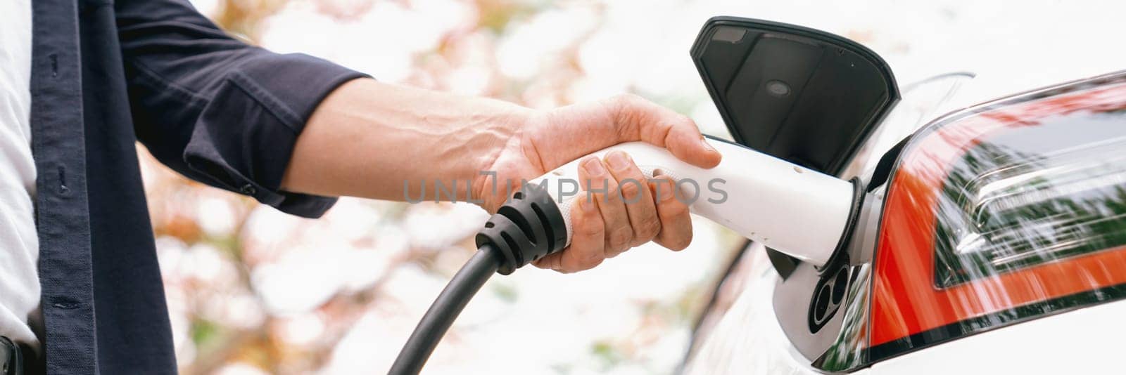 Panorama banner man recharging battery for electric car during autumnal road trip travel EV car in autumnal forest. Eco friendly travel on vacation during autumn with electric vehicle. Exalt