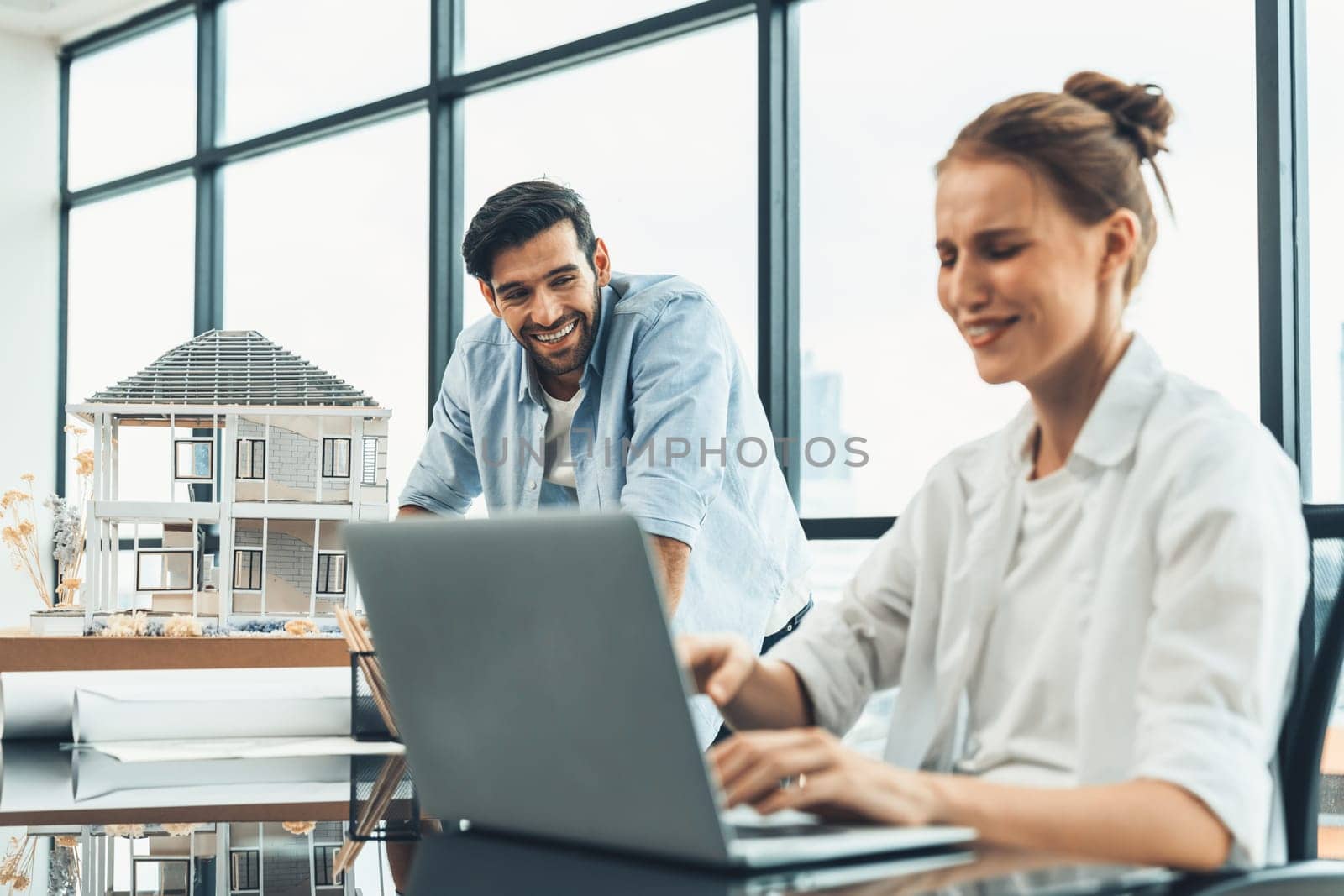 Young skilled project manager working by using laptop while caucasian engineer inspect house mode, using project plan at modern office or architect studio. Civil engineering, creative design. Tracery.