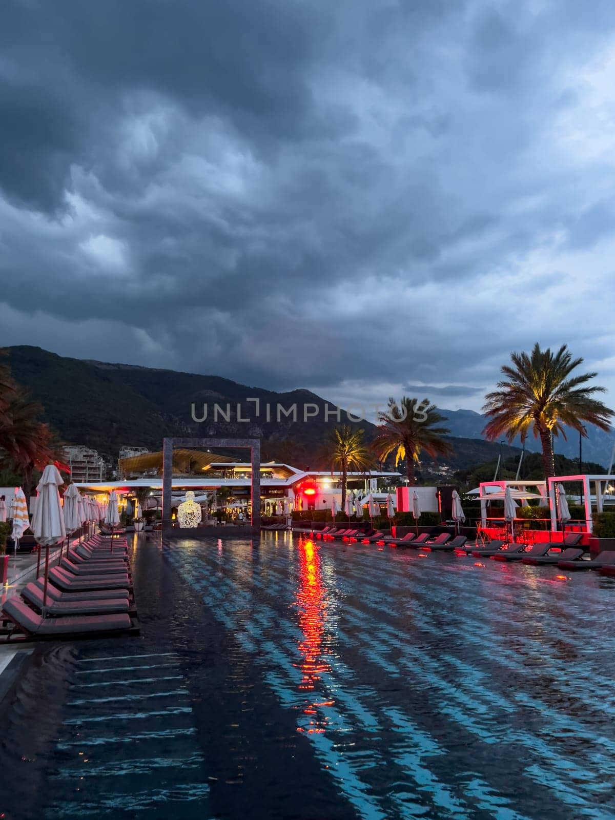 Sun loungers with folded umbrellas stand near the illuminated pool at dusk. High quality photo