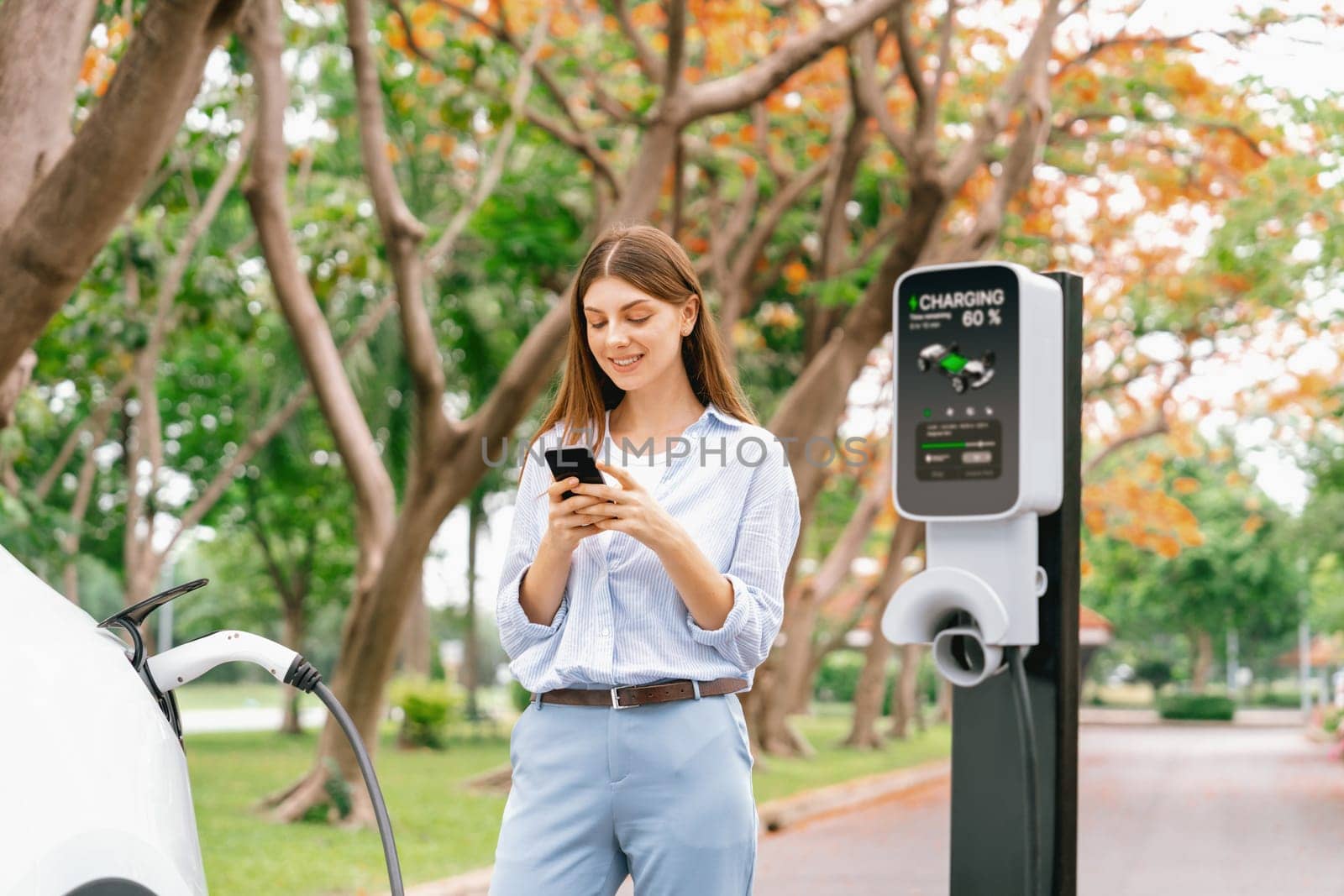 Young woman using smartphone online banking application to pay for electric car battery charging from EV charging station during autumn vacation holiday trip at national park or autumnal forest. Exalt