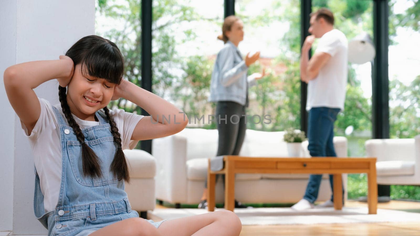 Stressed and unhappy girl huddle in corner, cover her ears with painful expression while her parent arguing in background. Domestic violence and traumatic childhood depression. Panorama Synchronos