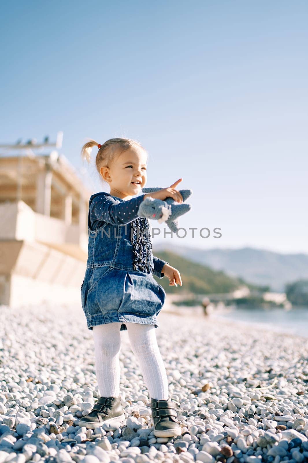 Little girl stands on the seashore and points into the distance with her hand with a toy. High quality photo