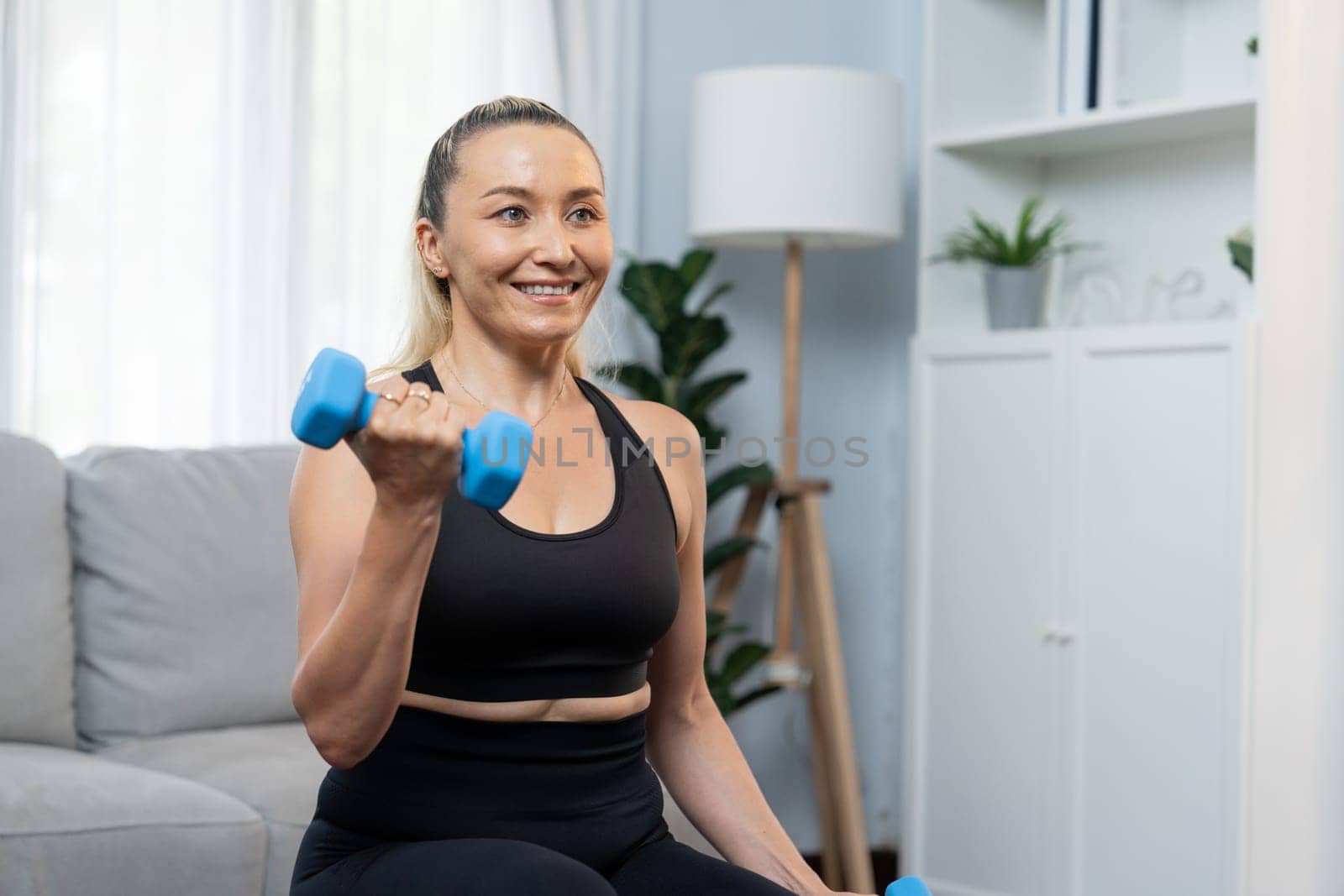 Athletic and sporty senior woman engaging in body workout routine, sitting on sofa and lifting dumbbell at home as concept of healthy fit body with body weight lifestyle after retirement. Clout