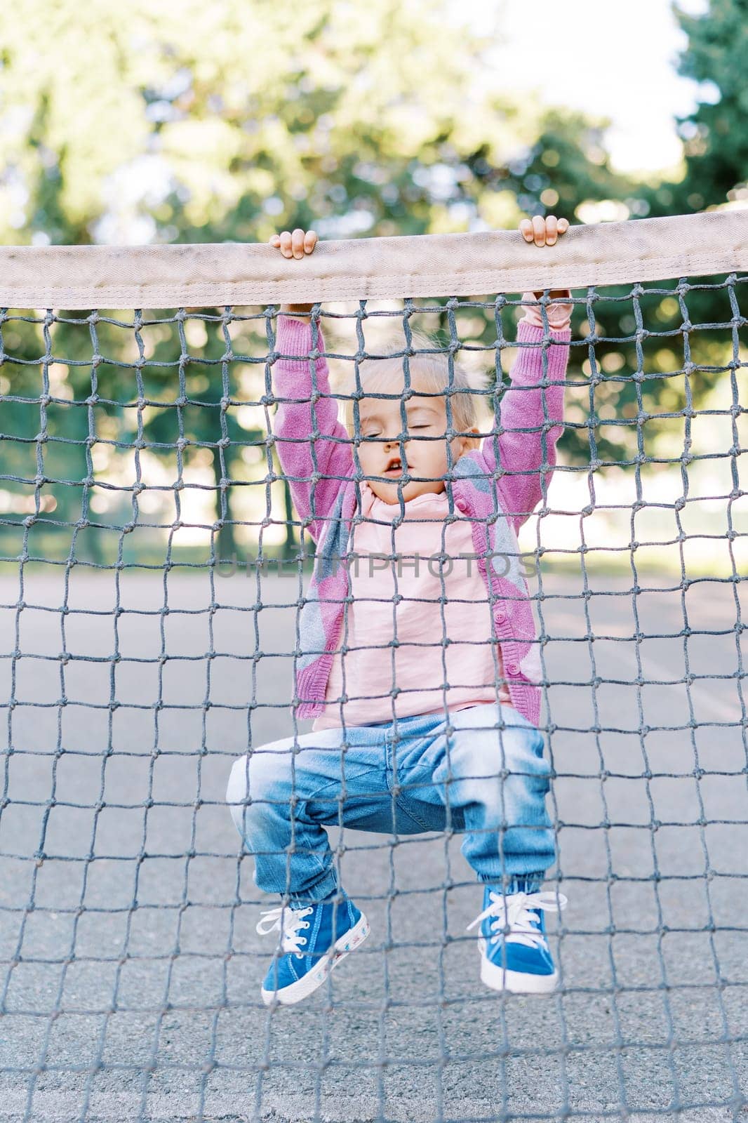 Little girl hangs on the edge of a tennis net with her legs folded. High quality photo