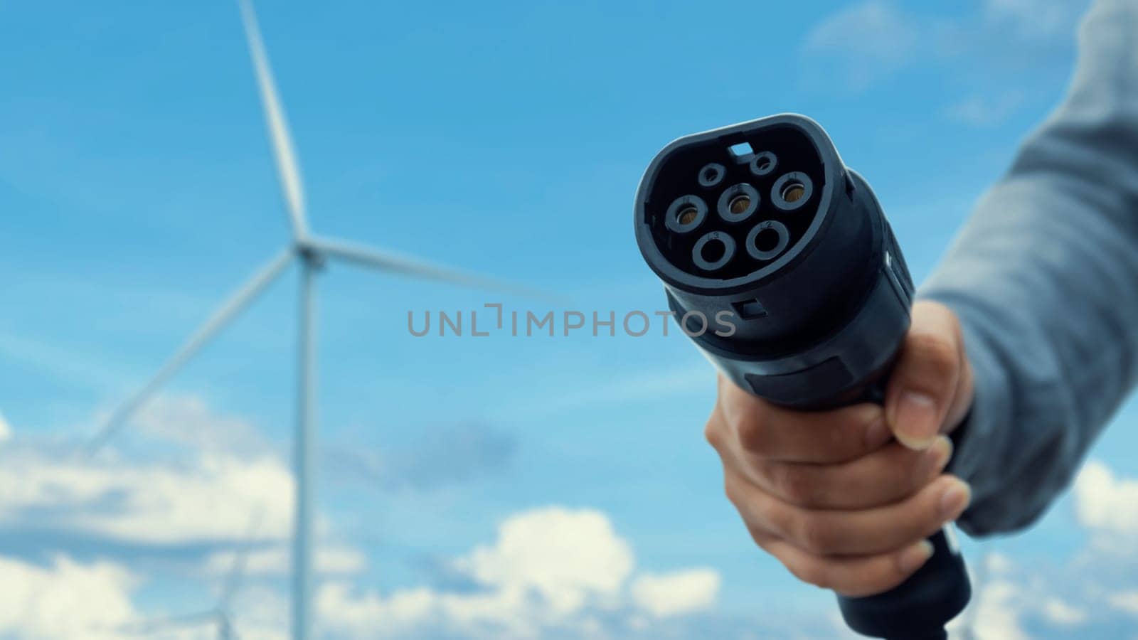 Focus EV charger pointing in front of camera with blurry businessman's hand wind turbine farm in background. Electric car charger plug using alternative clean energy reducing CO2 emission. Peruse