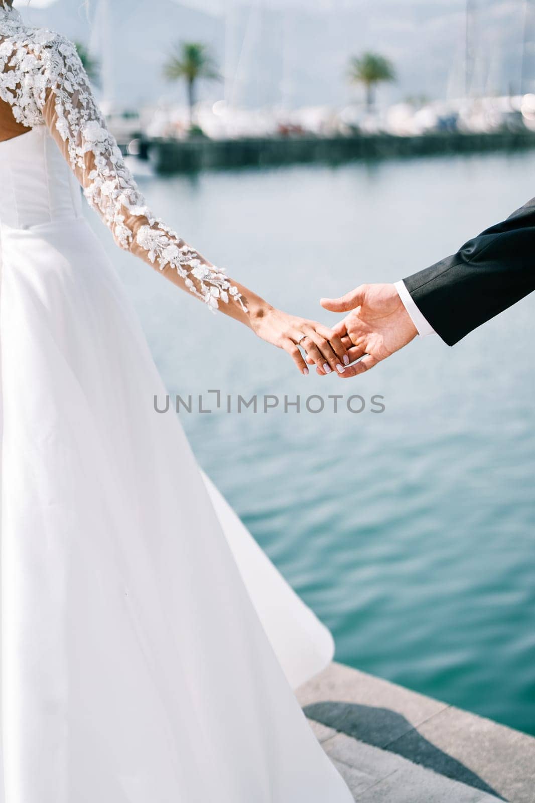 Bride hand touches the palm of groom standing on the pier. Cropped. Faceless by Nadtochiy