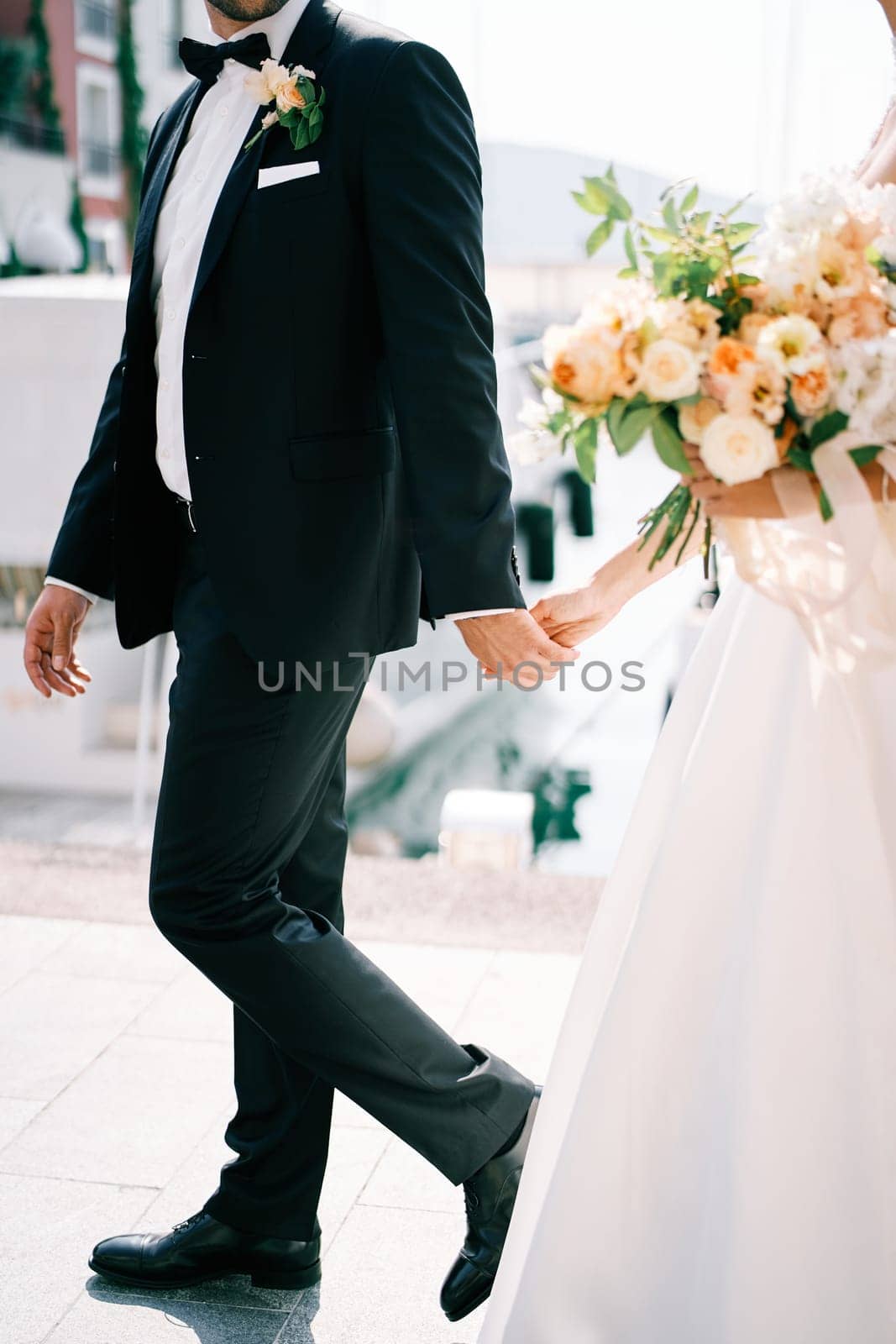 Bride and groom with a bouquet walk holding hands along the sea. Cropped. Faceless by Nadtochiy