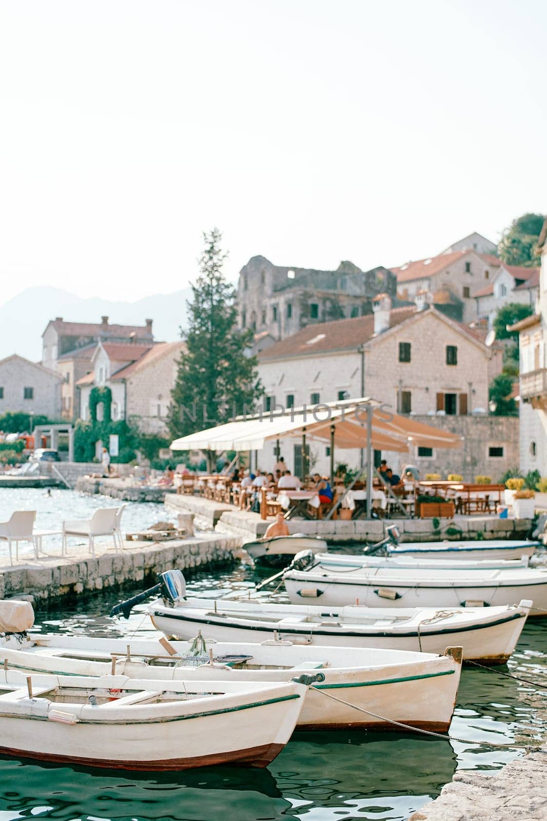 Fishing boats moored near the shore with a restaurant under a canopy near ancient houses. Perast, Montenegro. High quality photo