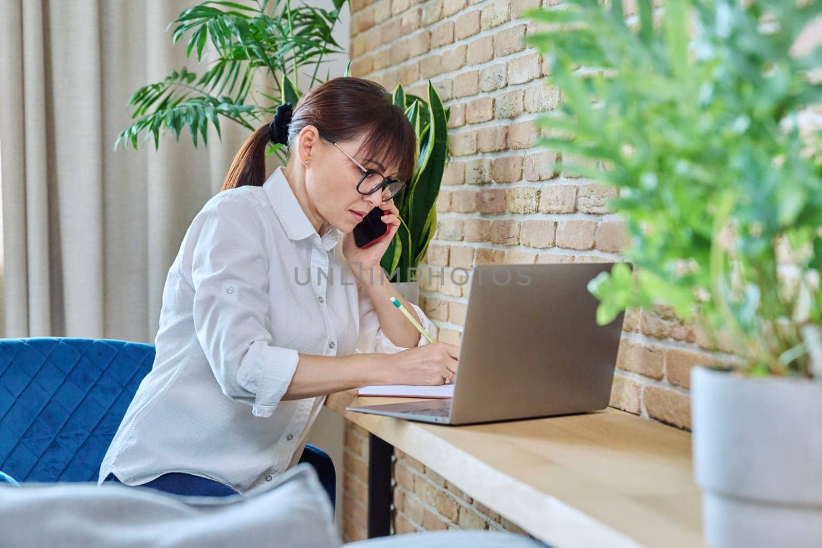 Middle-aged woman talking on mobile phone, sitting at desk at home with laptop computer. Female working remotely, freelancing, online services, technologies for work communication leisure