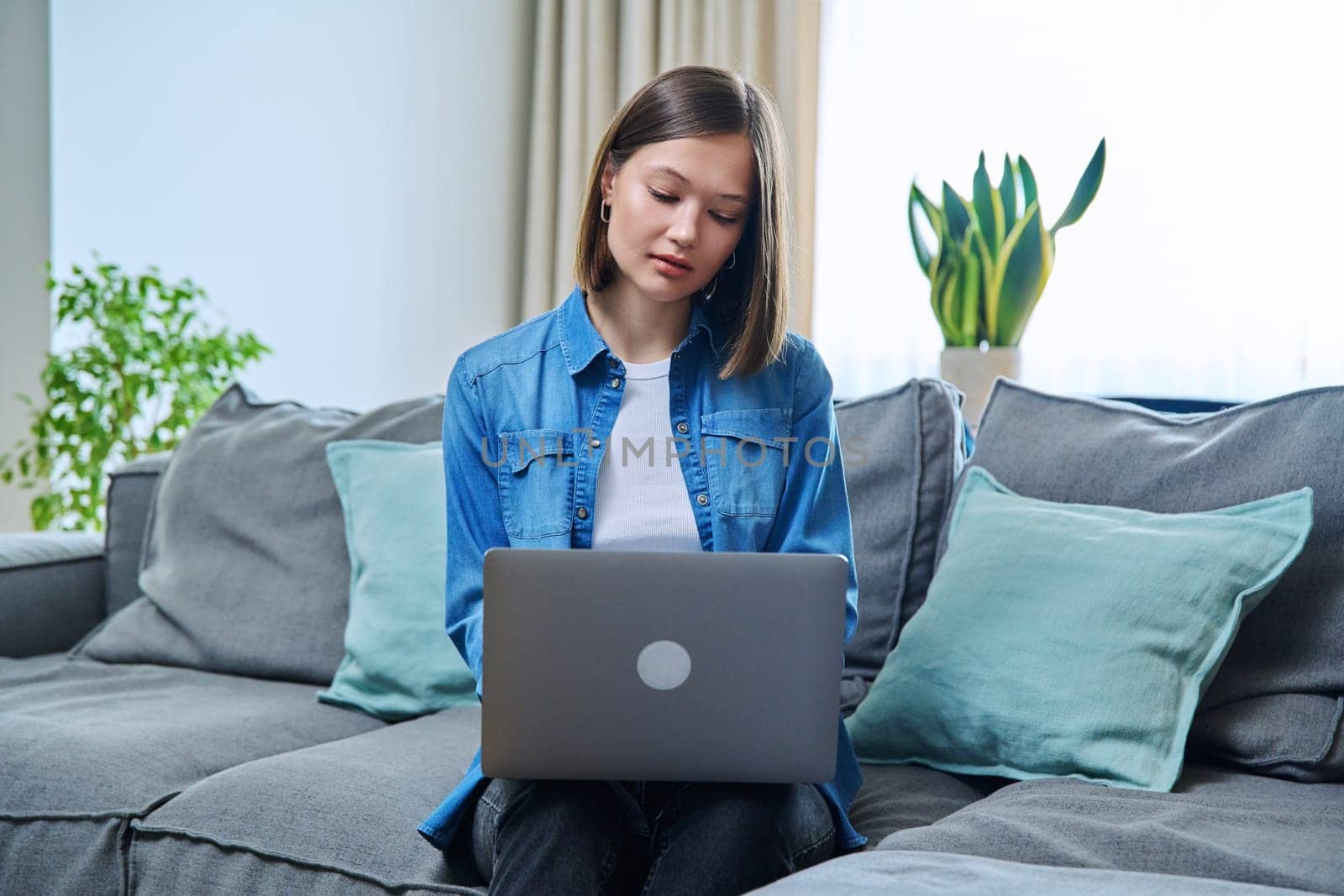 Young woman sitting on sofa in living room typing on laptop by VH-studio