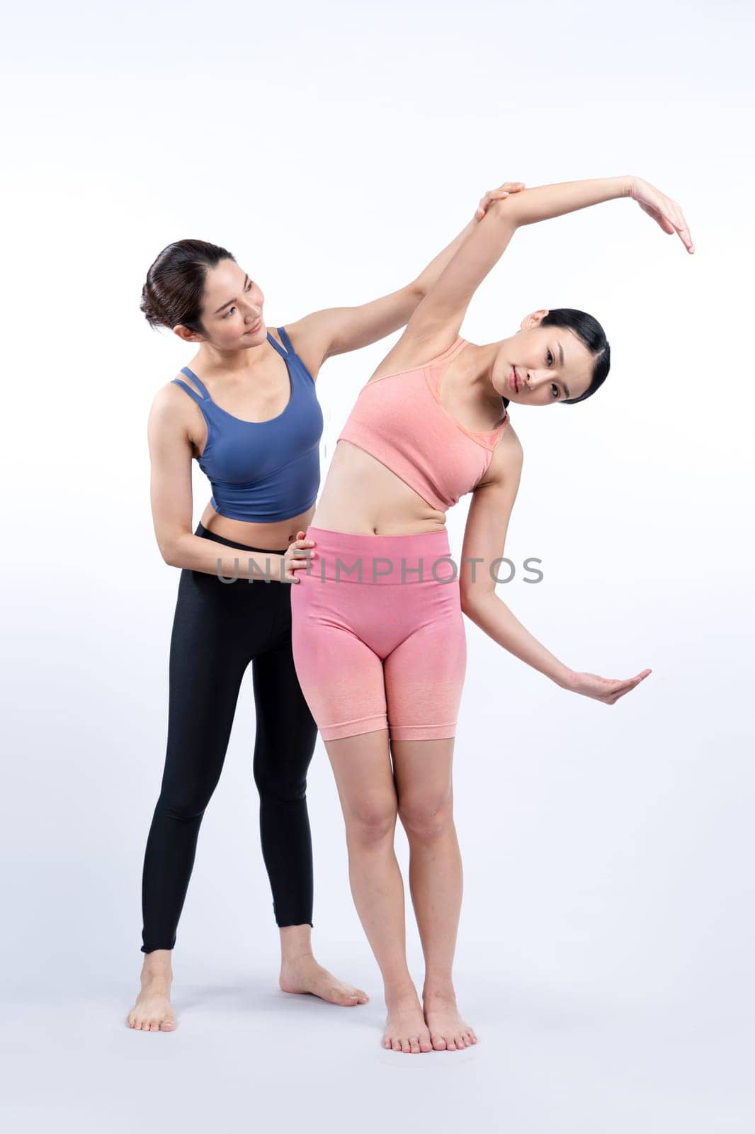 Asian woman in sportswear doing yoga exercise in standing pose with her trainer or yoga buddy. Healthy body care and meditation yoga lifestyle in full shot on isolated background. Vigorous