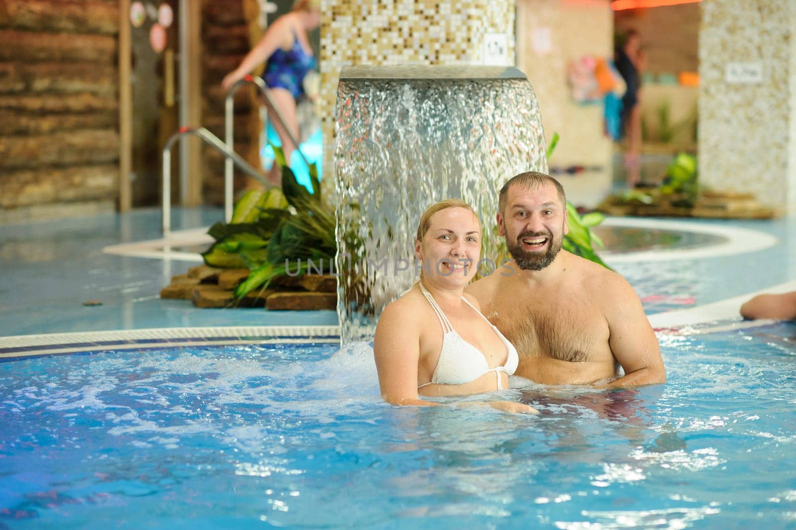 A man and woman enjoy the spray of a waterfall and swim in a pool.