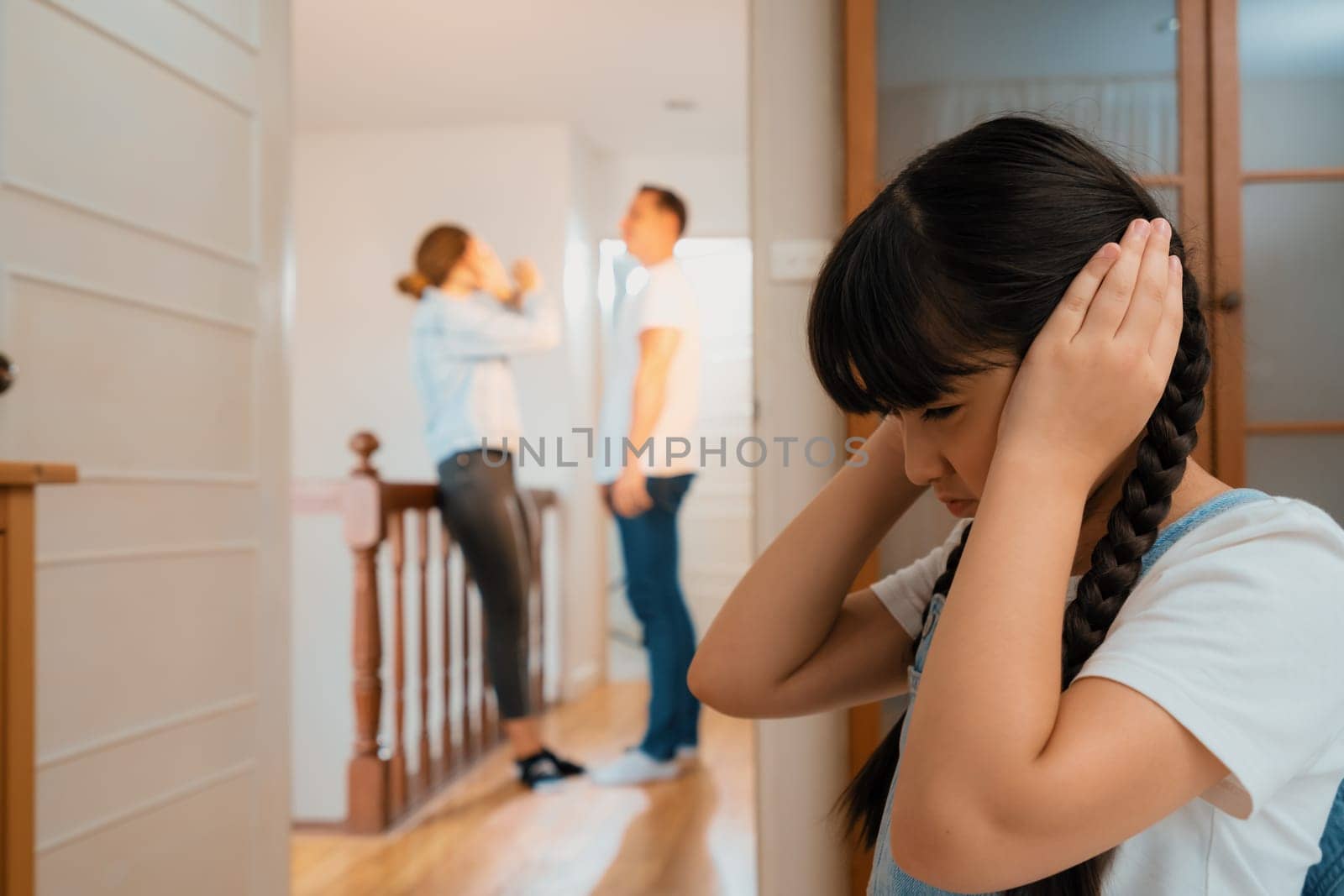 Stressed and unhappy young girl huddle in corner, cover her ears blocking sound of her parent arguing in background. Domestic violence at home and traumatic childhood develop to depression. Synchronos