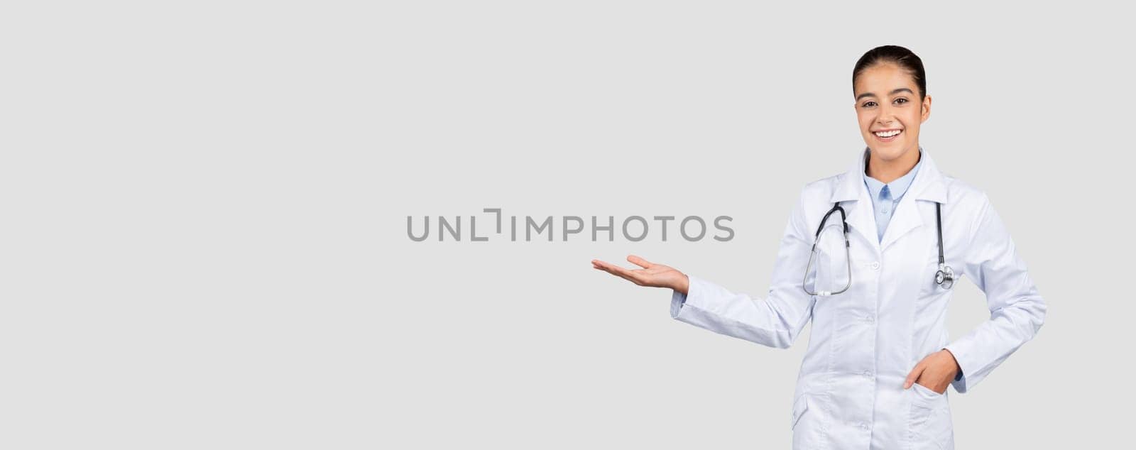 Cheerful european millennial doctor in white coat, with stethoscope hold copy space on hand, isolated on gray studio background, panorama. Medical ad and offer, health care presentation, sale
