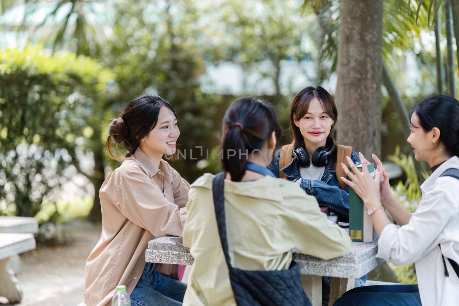 Group of young Asian college students sitting on a bench in a campus relaxation area, talking, sharing ideas, doing homework or tutoring for the exam together.