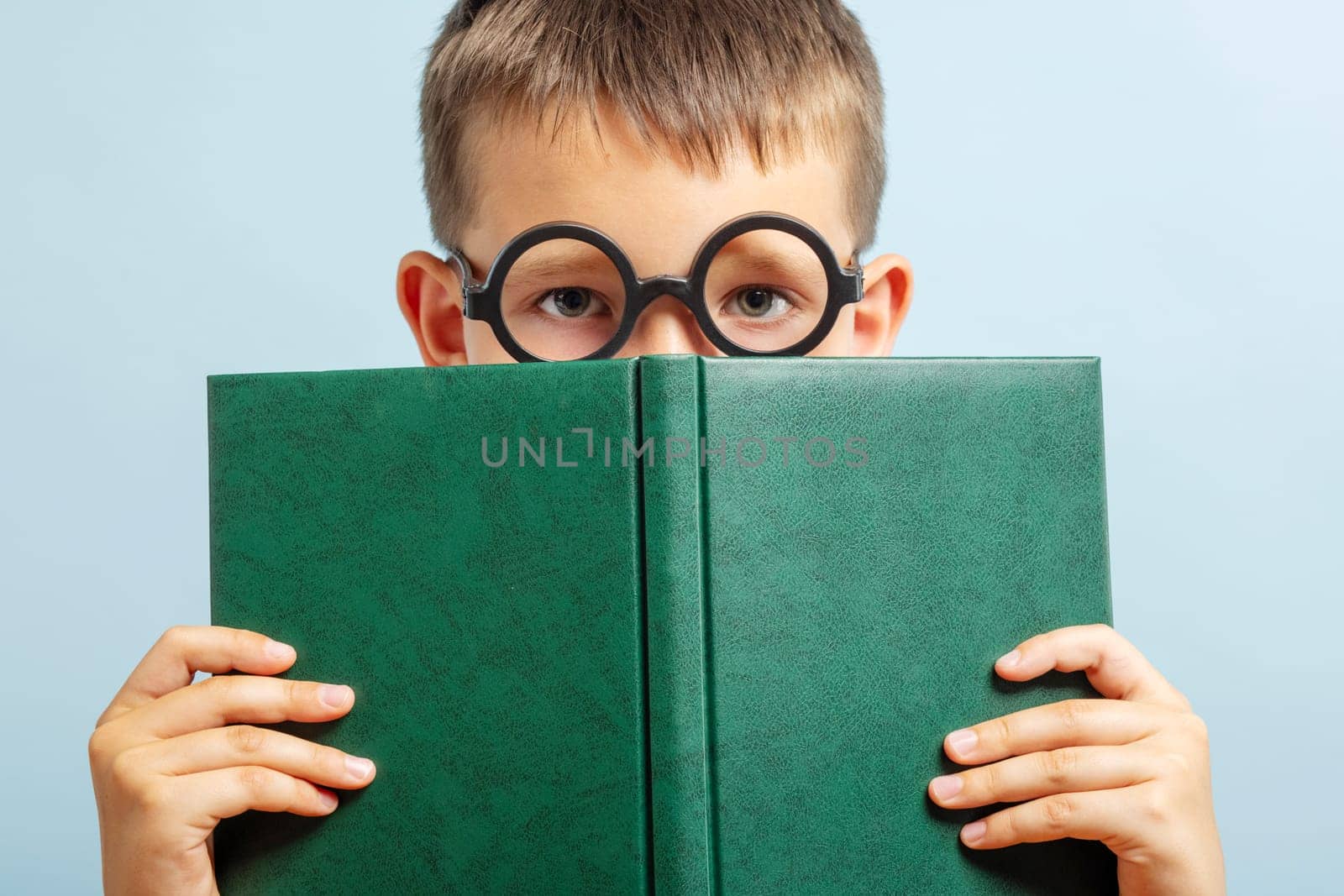 Little schoolboy in eyeglasses looks out from behind the book on blue background by andreyz
