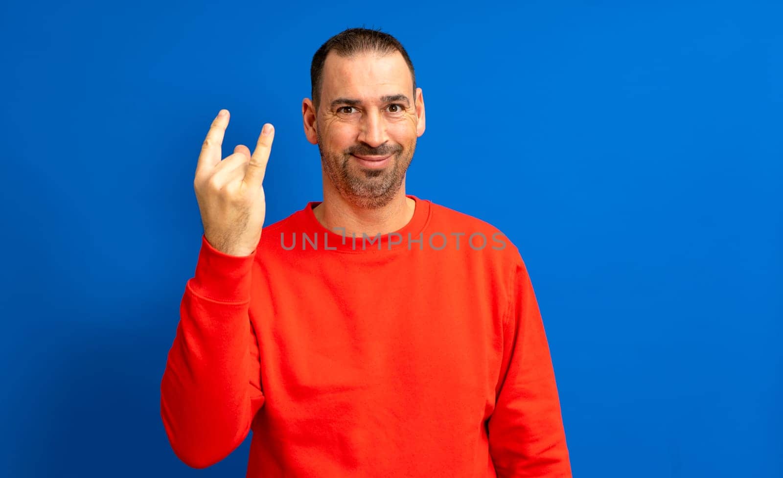 Hispanic man with beard in his 40s wearing a red sweatshirt doing rock gesture on blue studio background.