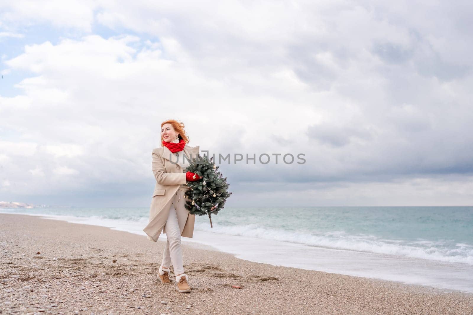 Redhead woman Christmas tree sea. Christmas portrait of a happy redhead woman walking along the beach and holding a Christmas tree in her hands. Dressed in a light coat, white suit and red mittens. by Matiunina