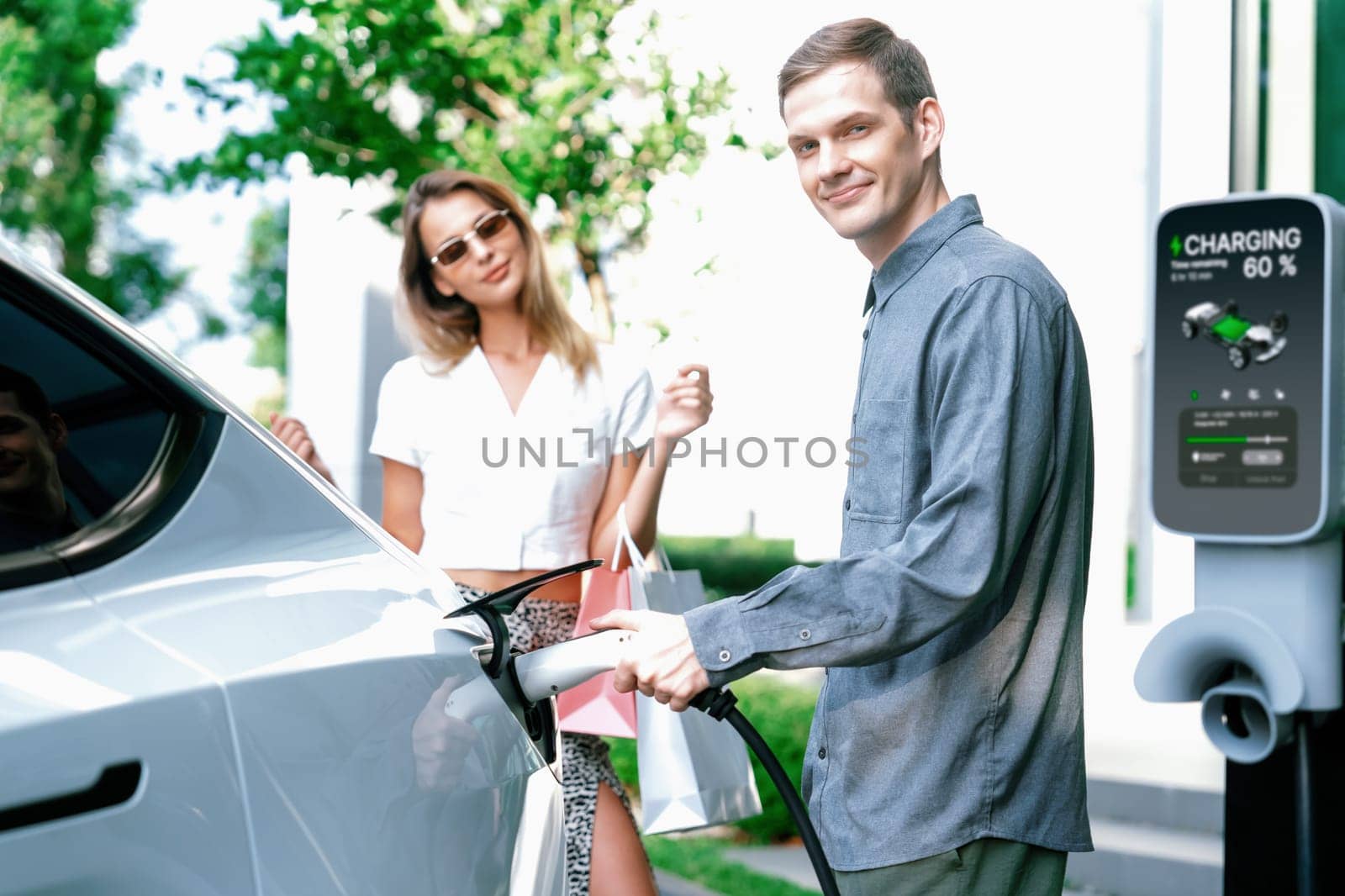 Young couple travel with EV electric car charging in green sustainable city outdoor garden in summer shows urban sustainability lifestyle by green clean rechargeable energy of electric vehicle innards