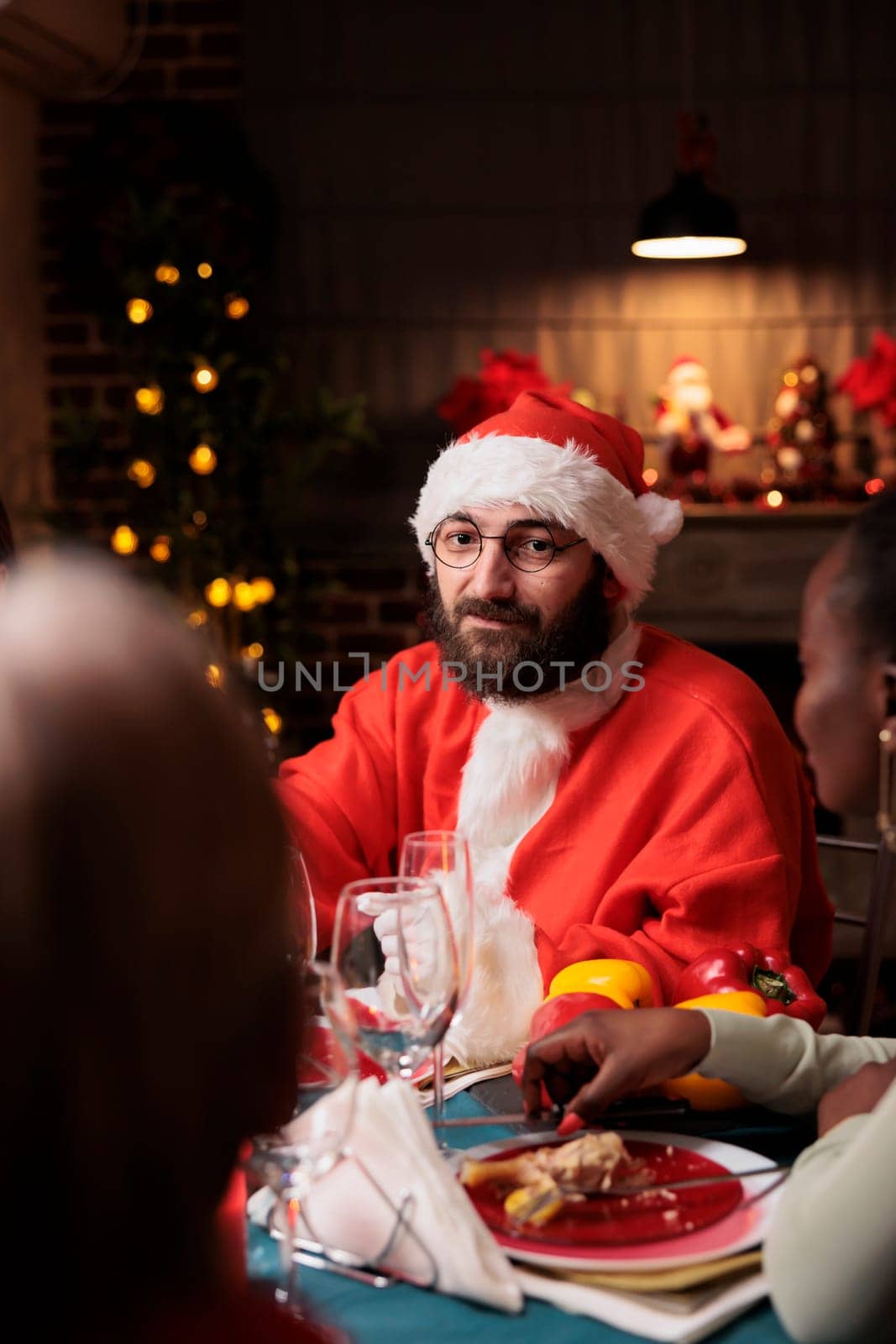 Santa claus man in costume sitting at homr with diverse friends and family, enjoying festive dinner on christmas eve holiday. Young adult with hat spreading positivity during december holiday.